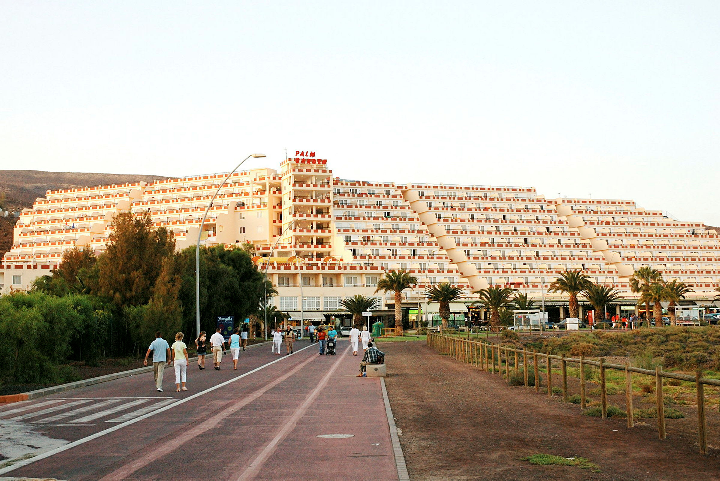 En stor bygning i pyramideform med flere etager ligger ved en promenade med mennesker, som spadserer og nyder omgivelserne. Træer og beplantning ses i forgrunden. (AI-genereret tekst)