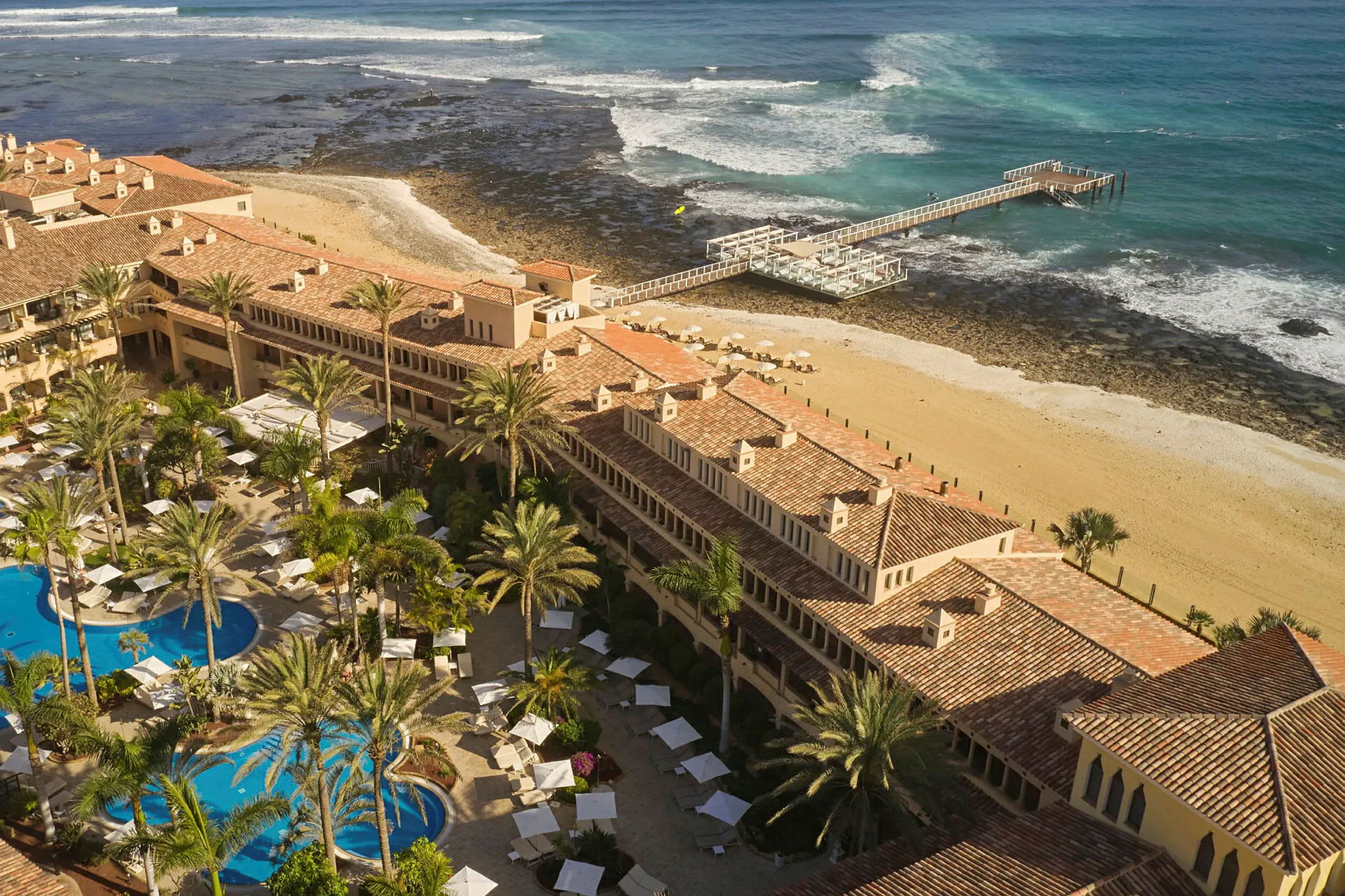 Luftfoto af et feriested med pools, liggestole og palmer tæt på en strand og mole ved havet. (AI-genereret tekst)