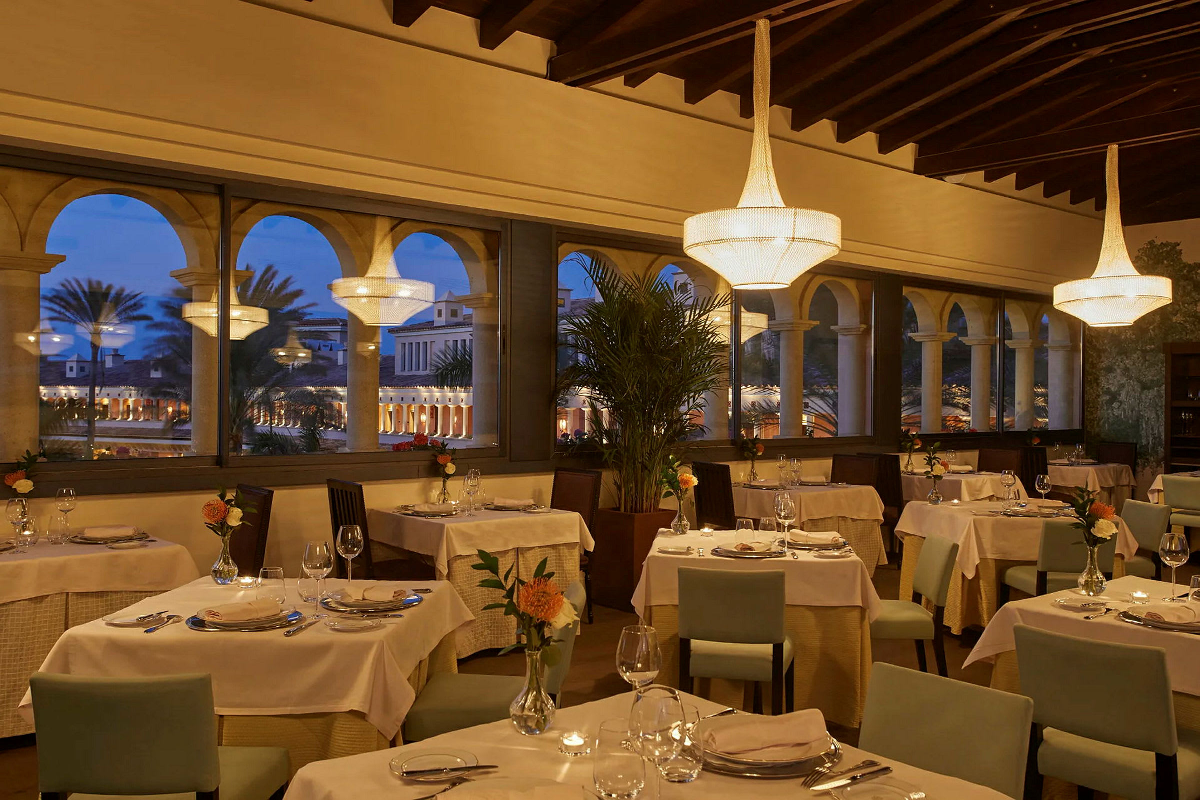 Interiør af en elegant restaurant med opdækkede borde og store vinduer med udsigt til en oplyst indre gård om aftenen. (AI-genereret tekst)