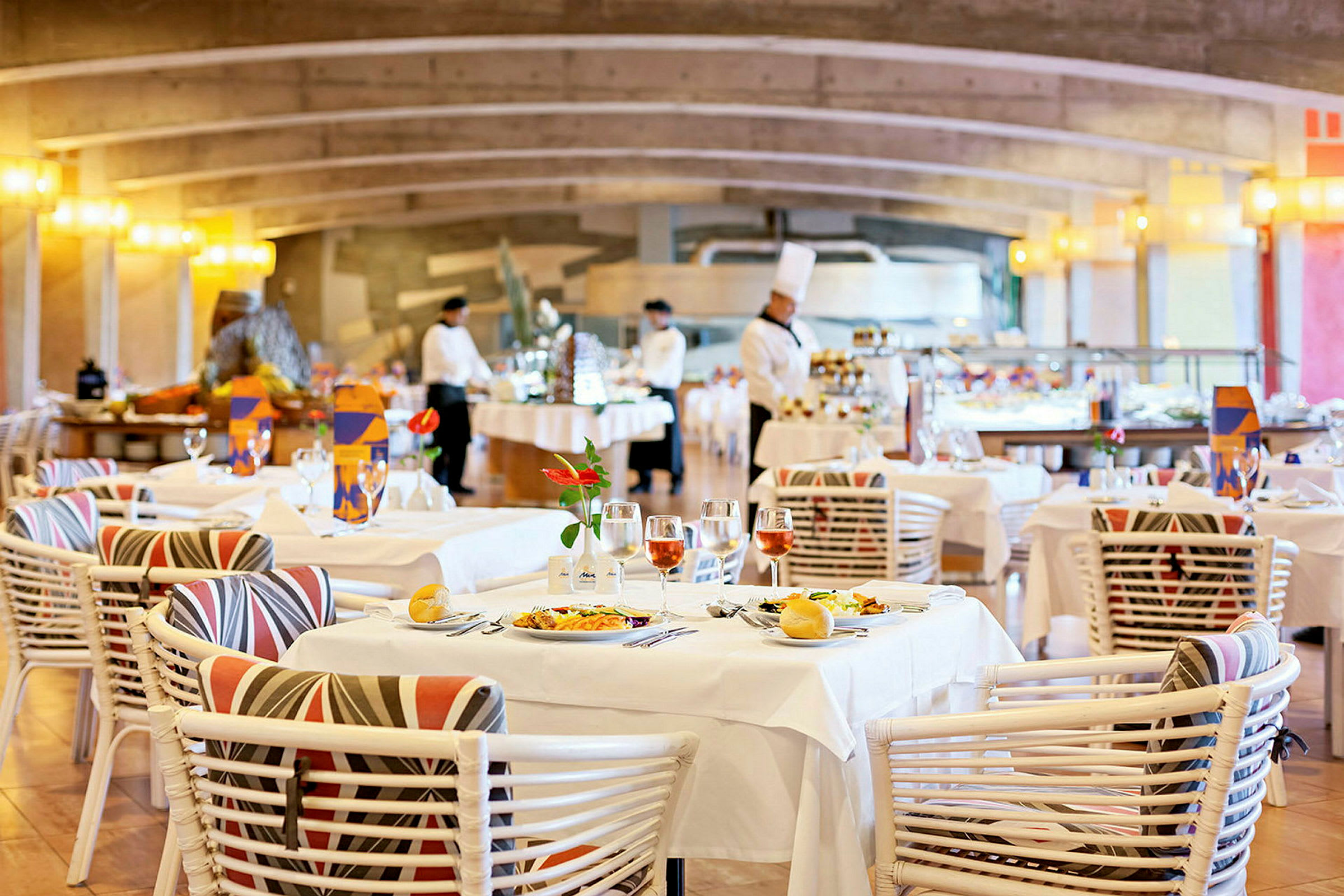 Et restaurantområde med dækkede borde, dekoreret med blomster og vinglas. I baggrunden ses personalet i arbejde ved buffeten. (AI-genereret tekst)