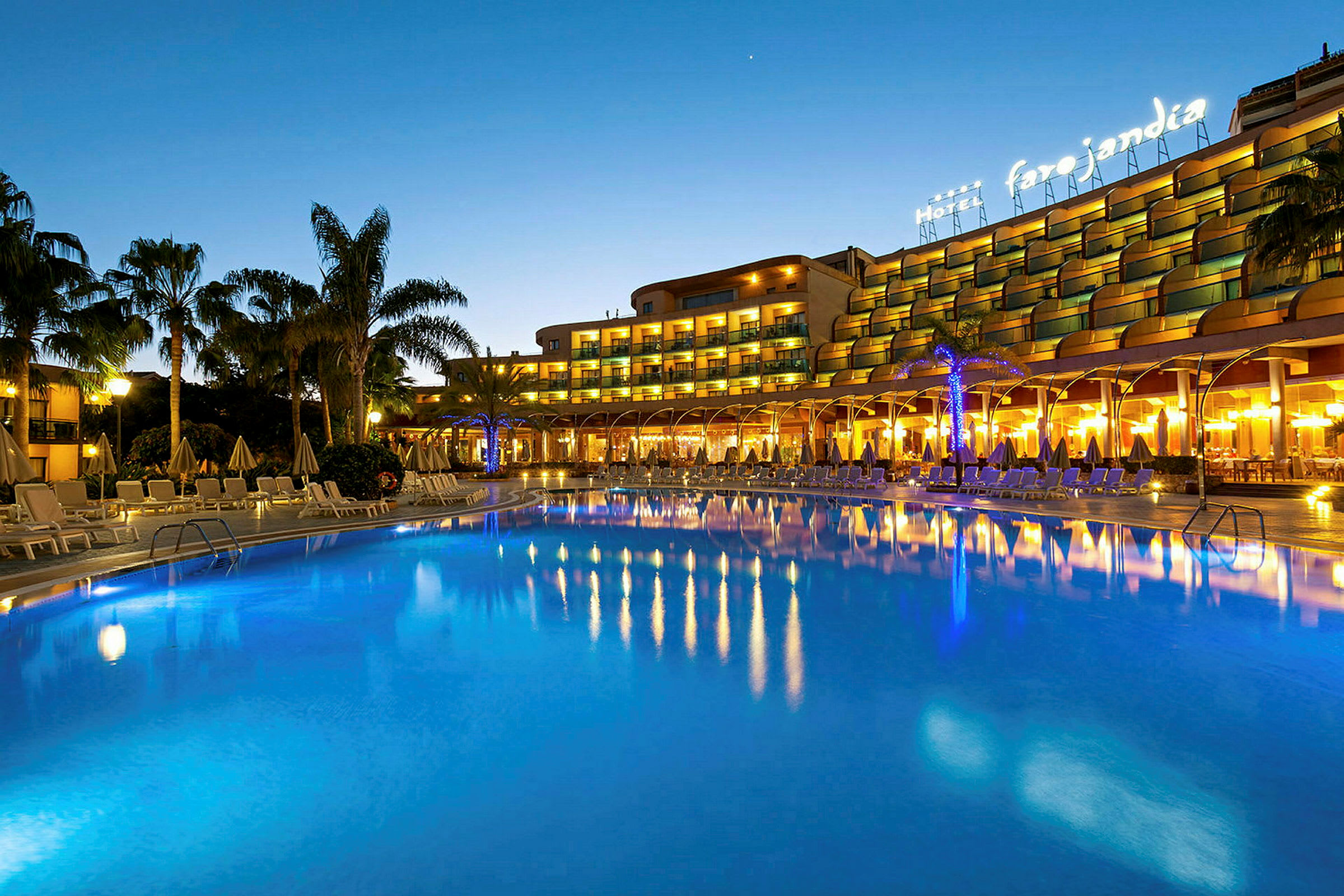 Oplyst hotel ved en stor opvarmet pool omgivet af liggestole og palmer om aftenen. (AI-genereret tekst)