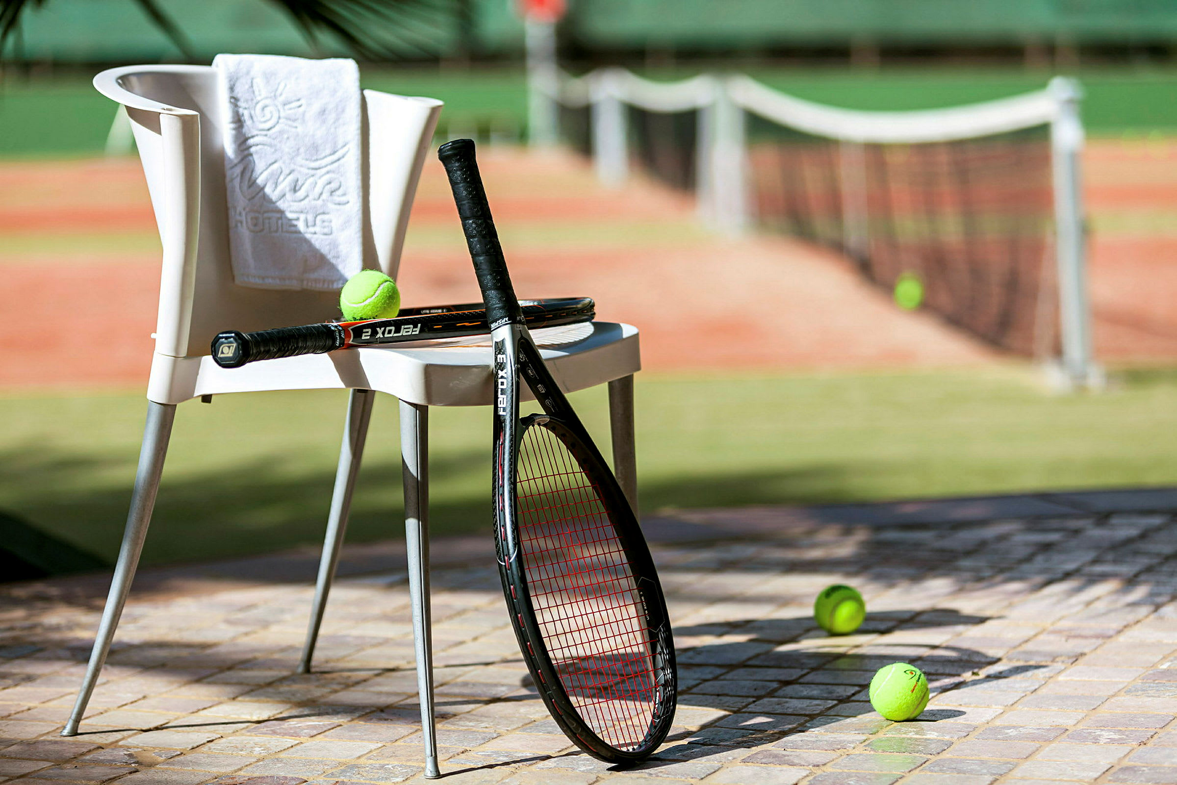 En stol med et håndklæde og to tennisracketer placeret ved siden af en tennisbane, med tennisbolde på jorden. (AI-genereret tekst)