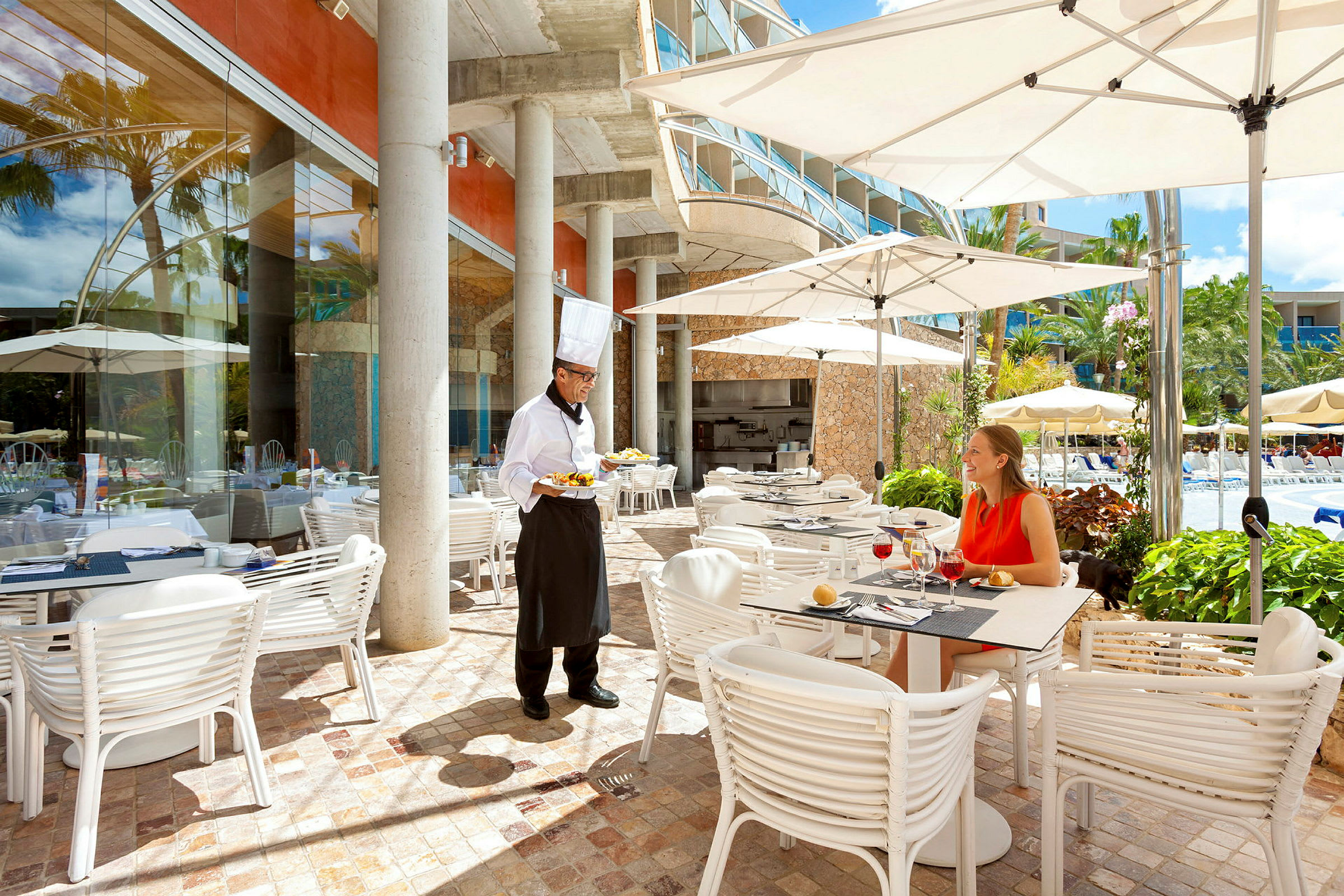 En restaurantterrasse med borde, stole og parasoller ved siden af en pool. En tjener serverer mad til en gæst. (AI-genereret tekst)