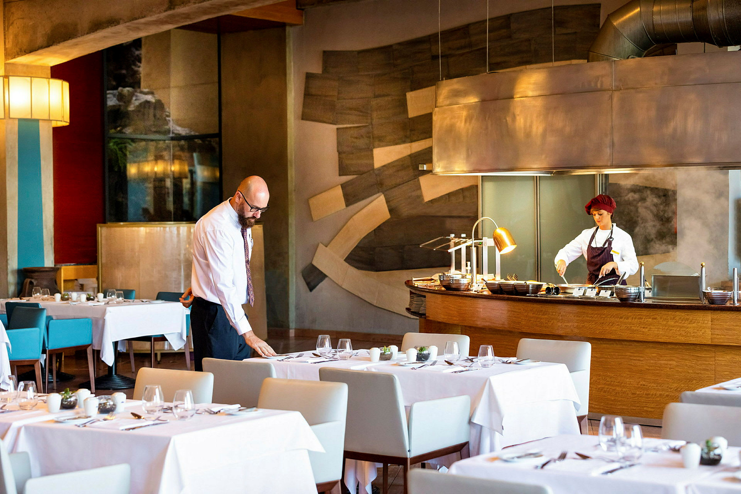 Restaurantinteriør med dækkede borde og en køkkenstation med en person i arbejdstøj. (AI-genereret tekst)