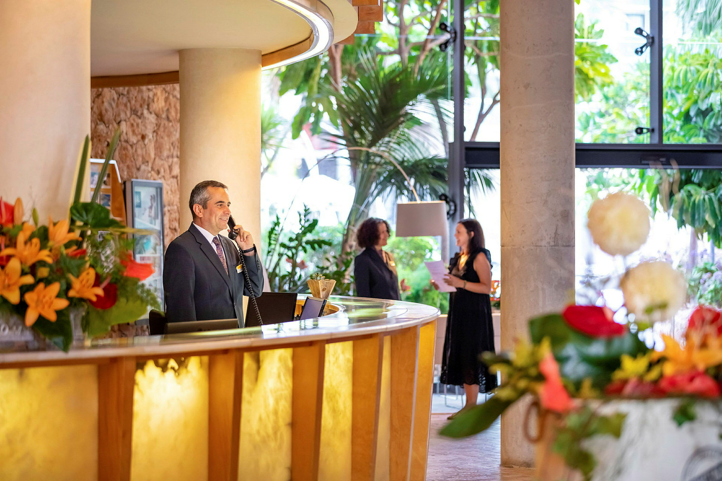 En hotelreception med blomster, en receptionist ved skranken og gæster i baggrunden. (AI-genereret tekst)