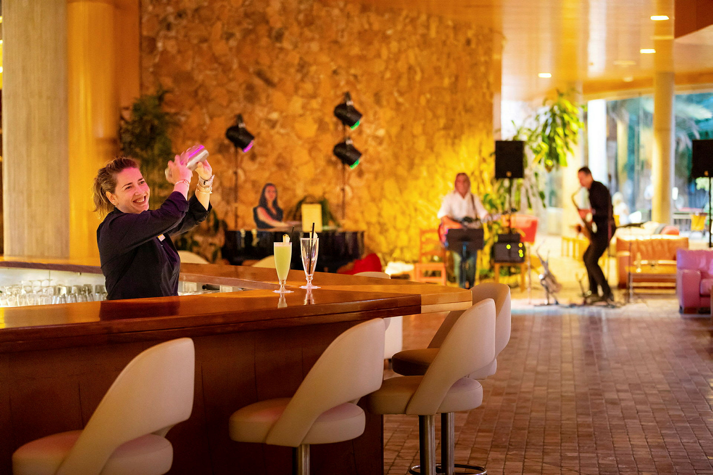 En bartender laver en drink ved bardisken. I baggrunden ses musikere spille instrumenter i en varm og stilfuld atmosfære. (AI-genereret tekst)