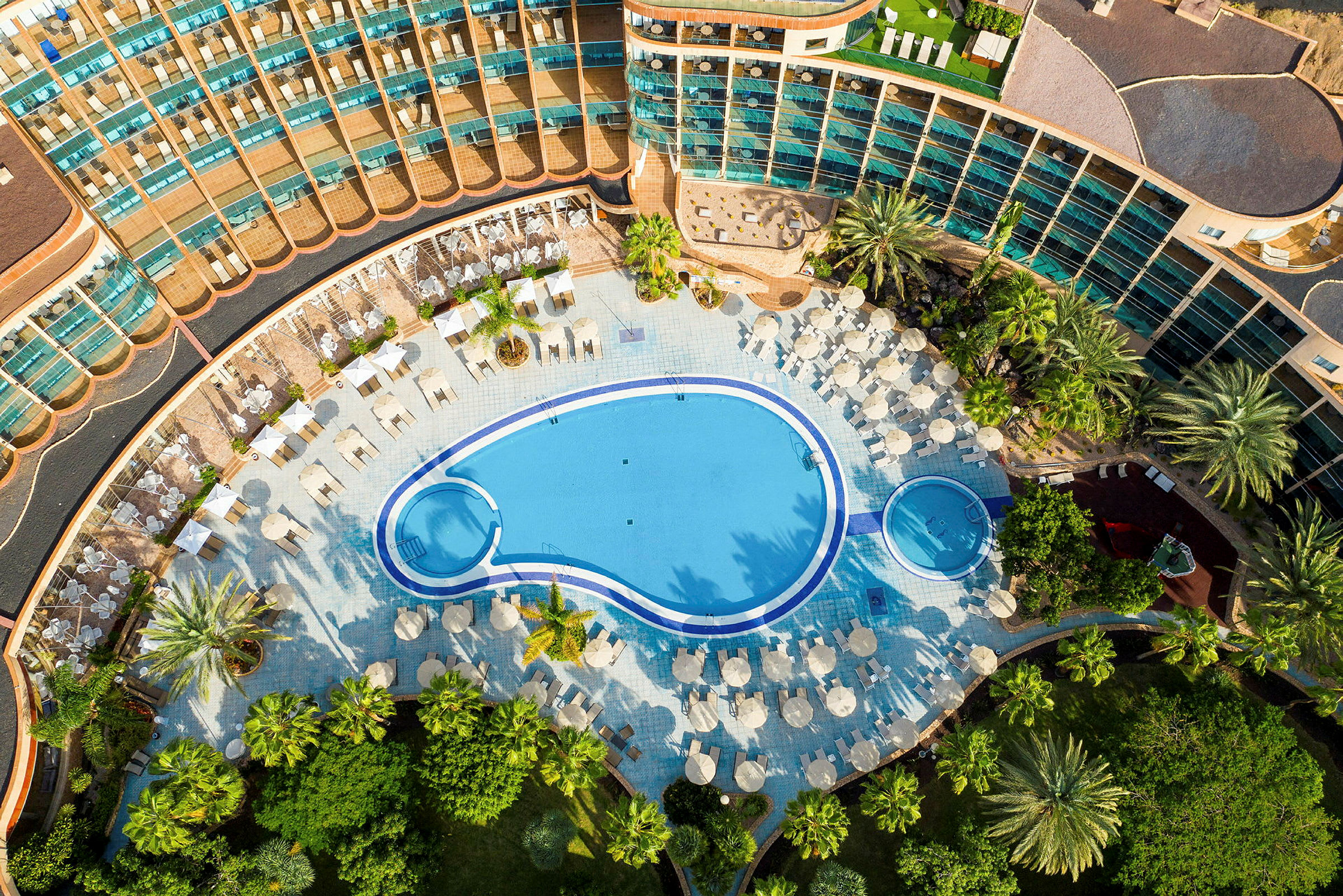 Luftfoto af et hotelområde med swimmingpool og liggestole, omgivet af frodige haver. (AI-genereret tekst)