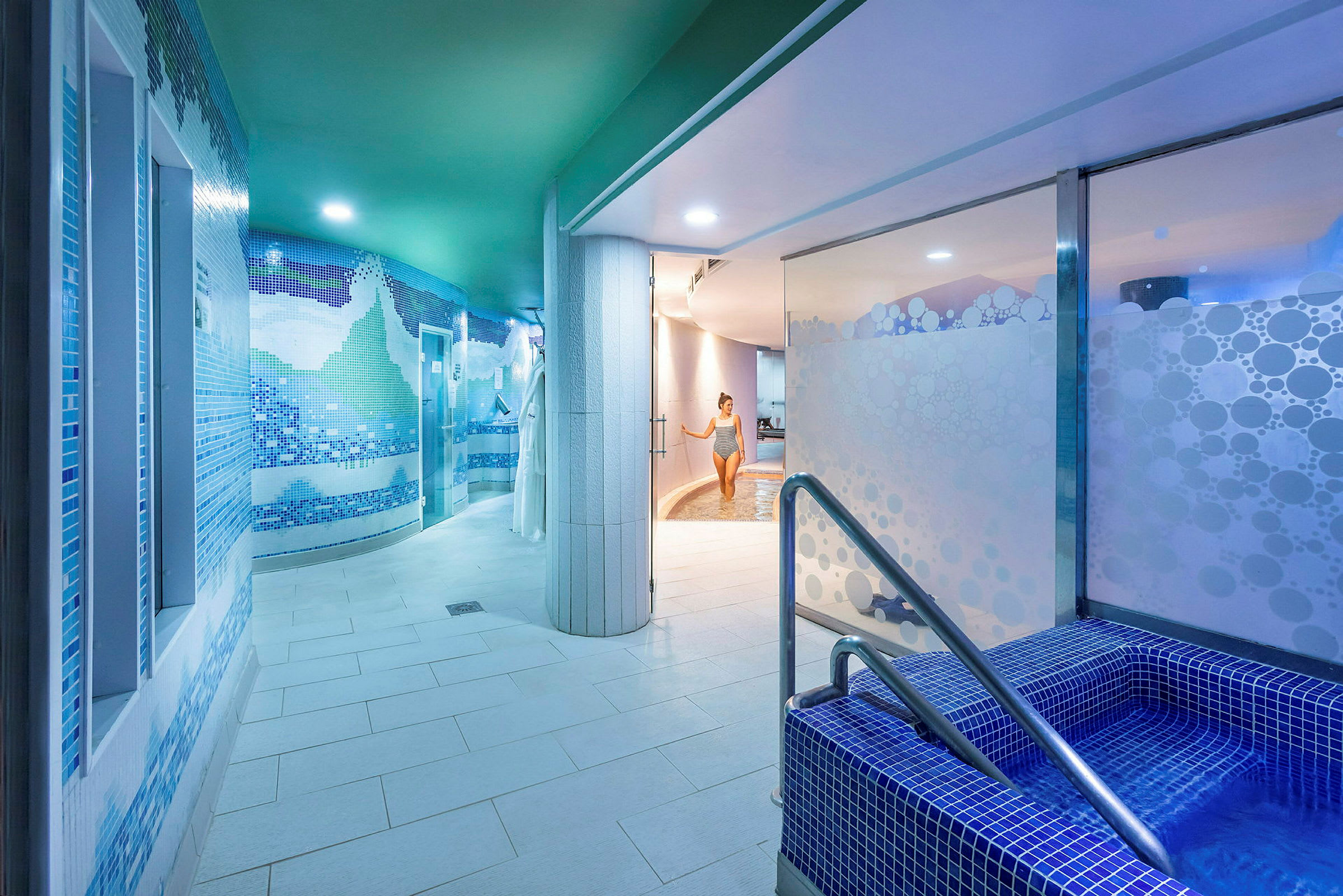 Et indendørs spa med blå mosaikvægge, en mindre pool med trapper i forgrunden og en person, der går mod en større pool i baggrunden. (AI-genereret tekst)