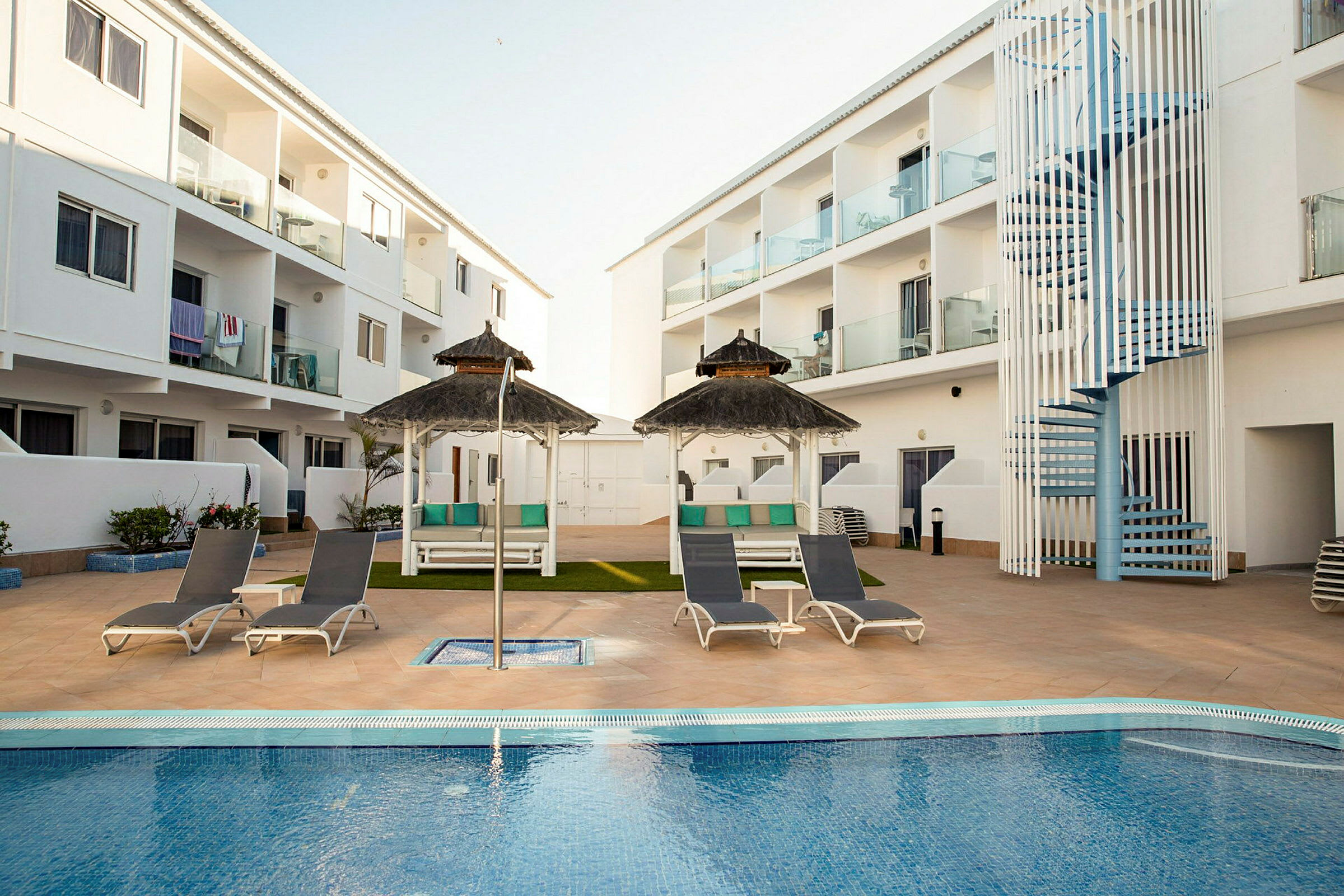 Et hotelområde med en swimmingpool omgivet af liggestole og parasoller. I baggrunden ses hvide bygninger med altaner. (AI-genereret tekst)