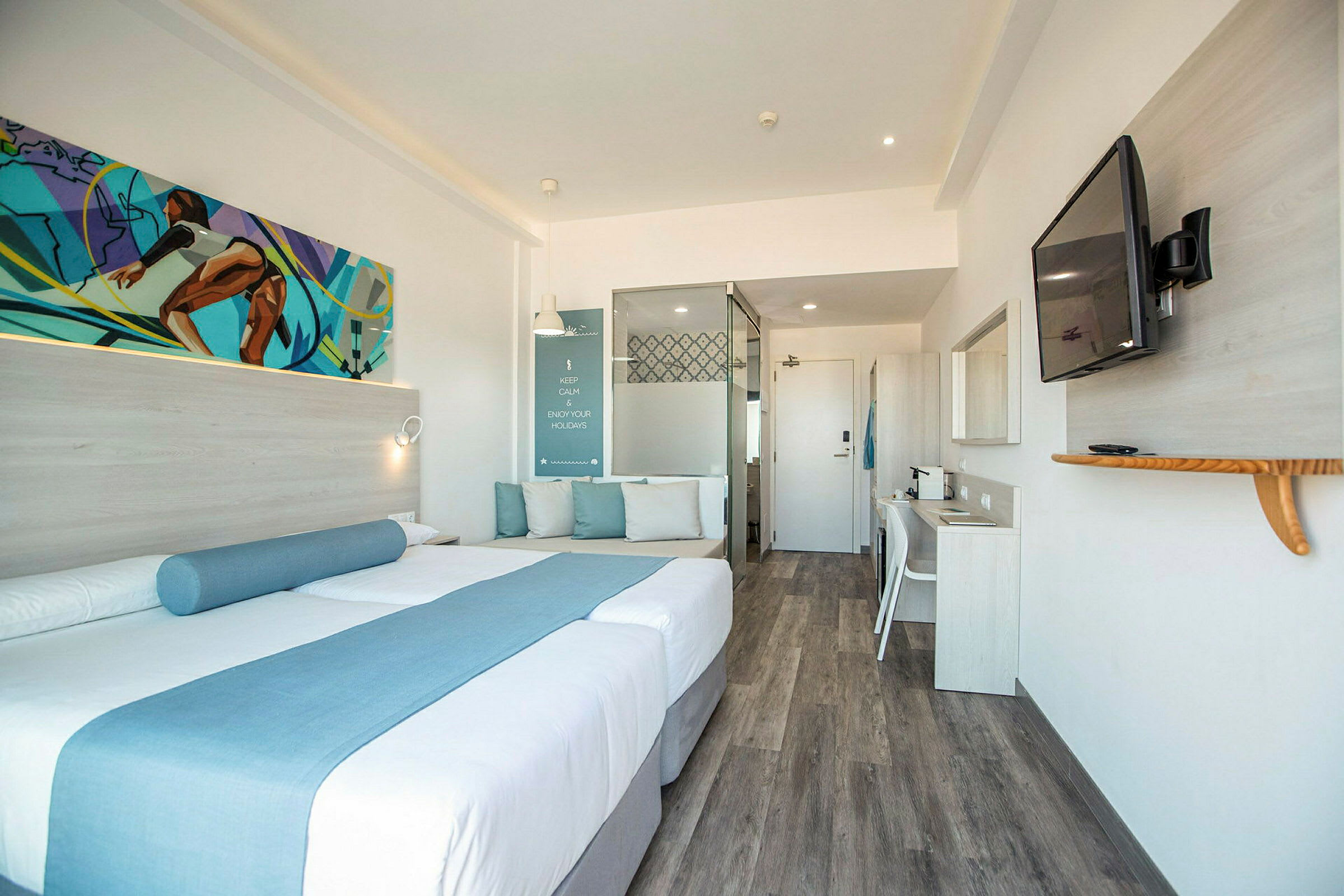 Et moderne hotelværelse med en dobbeltseng dekoreret med blå tekstiler, en siddeplads med puder, et skrivebord og et vægmonteret fjernsyn. (AI-genereret tekst)