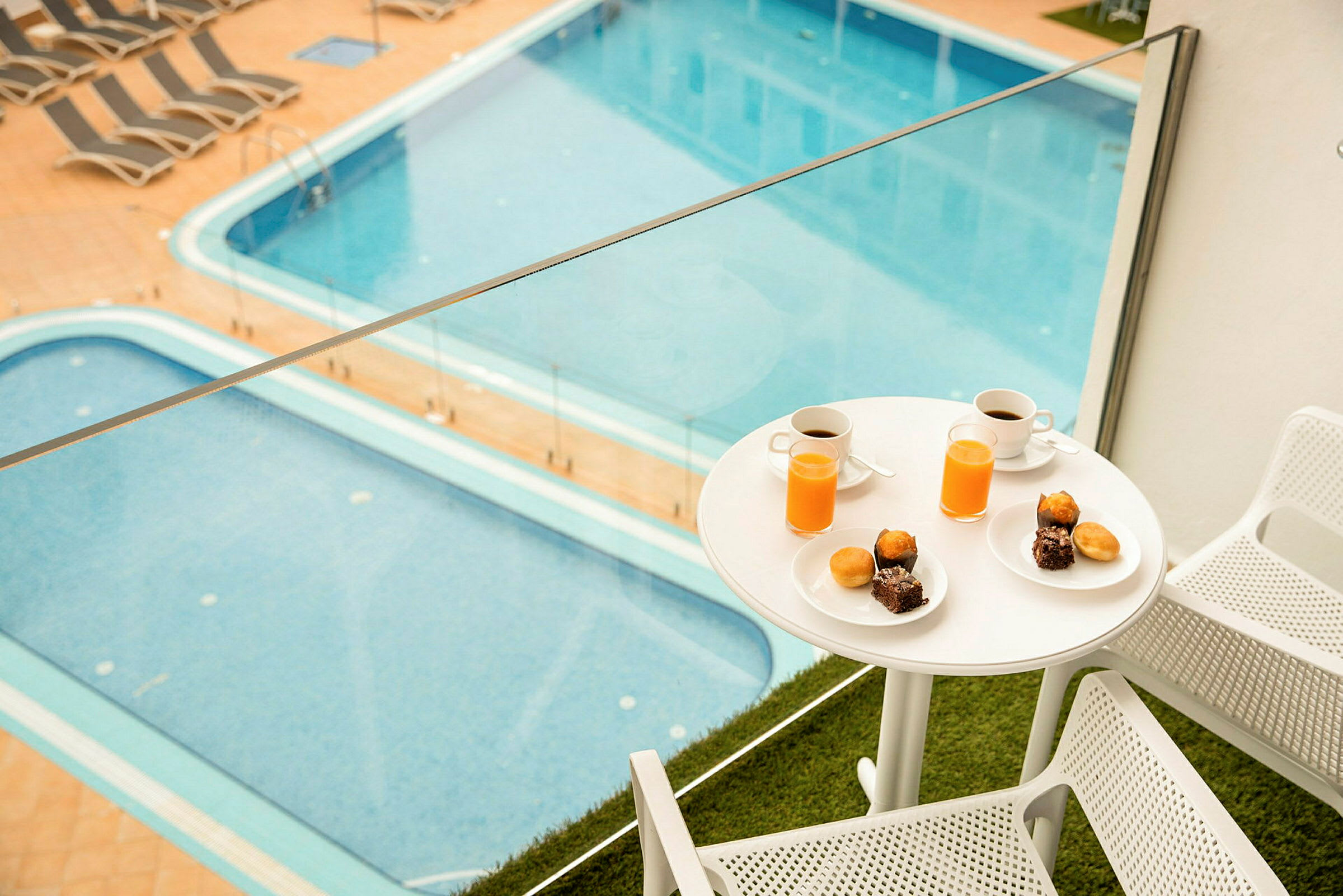 Billede af en altan med to stole og et bord med morgenmad, inklusive kaffe og juice, samt udsigt over en swimmingpool. (AI-genereret tekst)