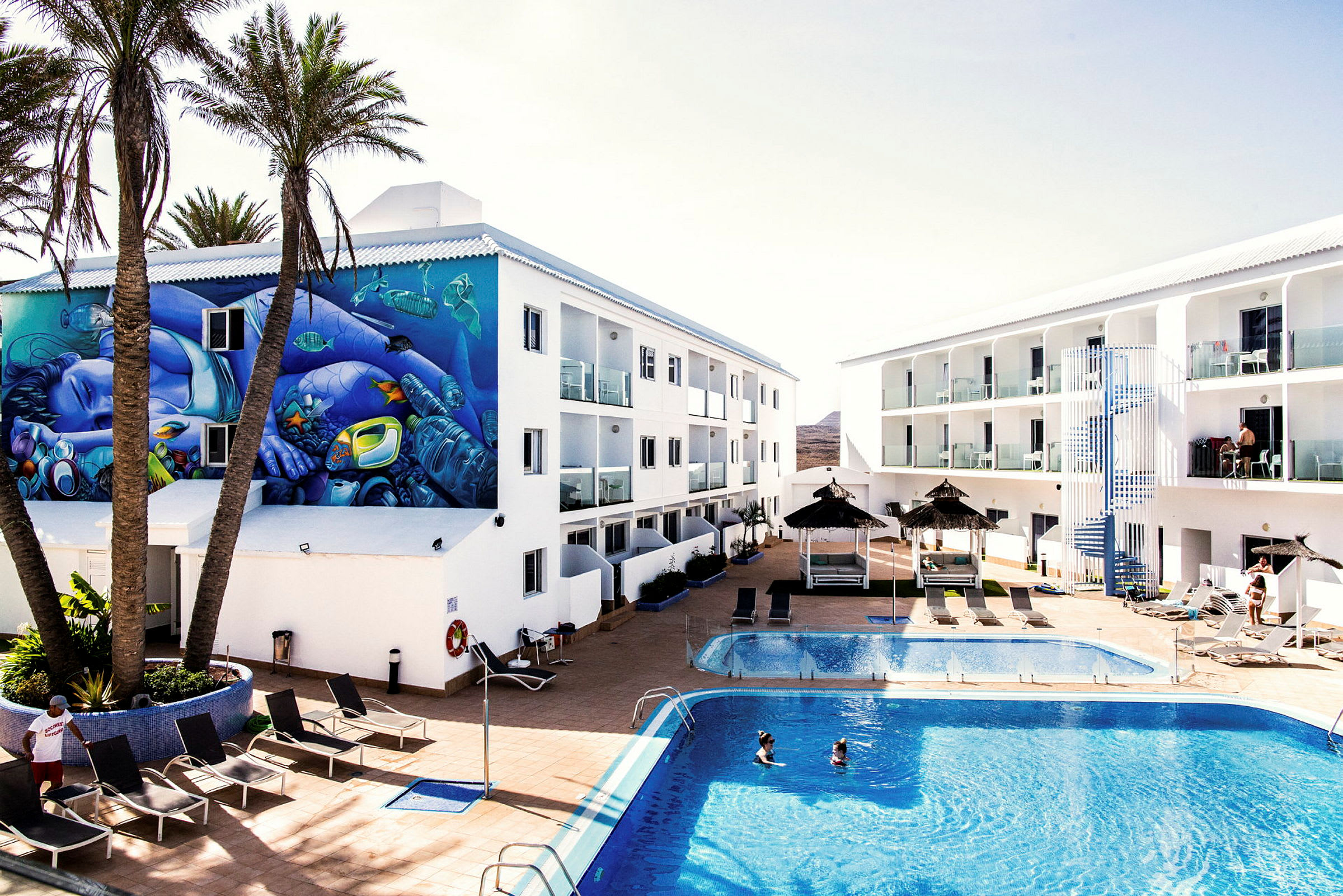 Hotelområde med pool, liggestole, parasoller og to hvide bygninger. En mur har et farverigt vægmaleri, og to personer svømmer i poolen. (AI-genereret tekst)