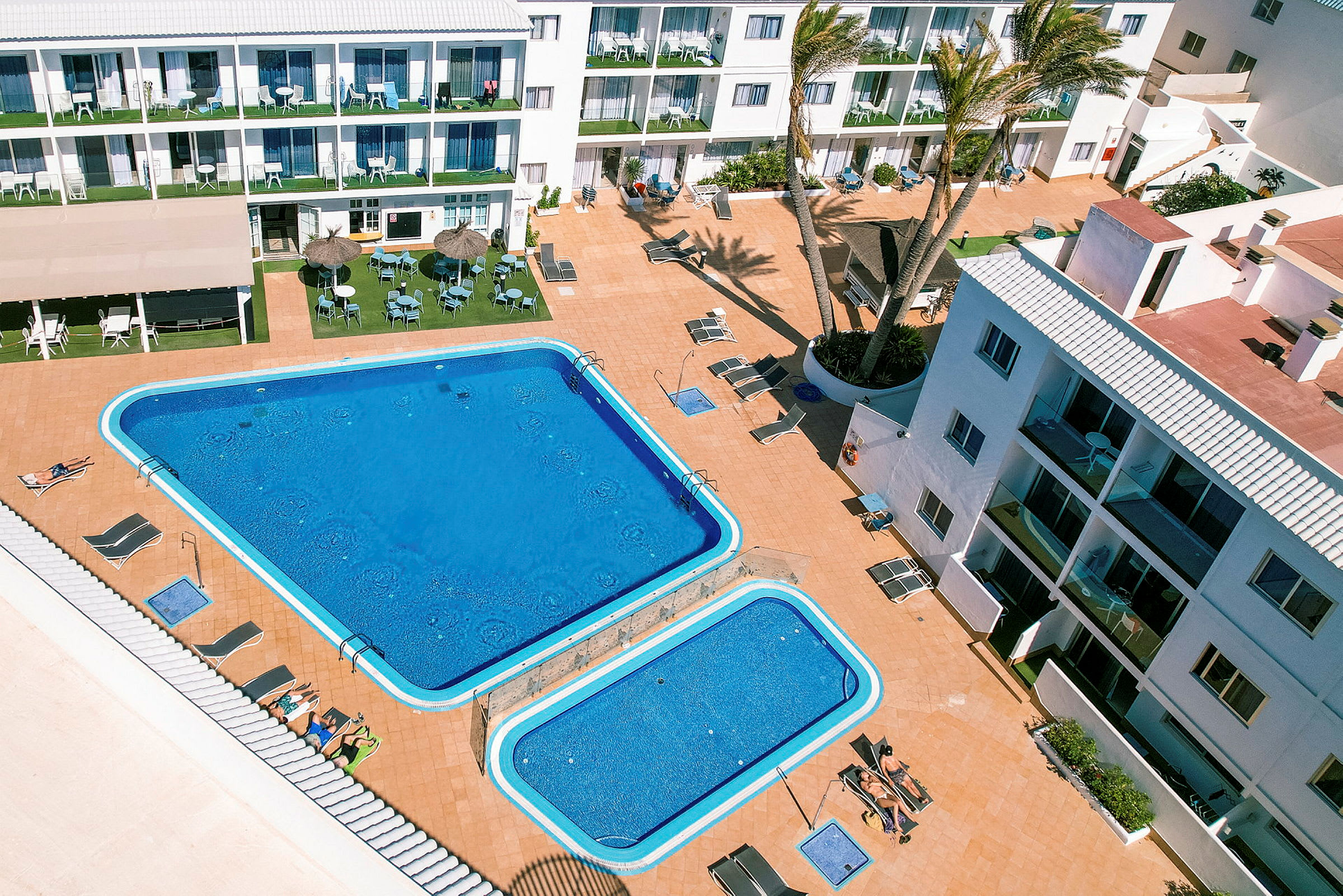 Billede af et hotelområde med to swimmingpools samt stole og parasoller på terrassen. (AI-genereret tekst)