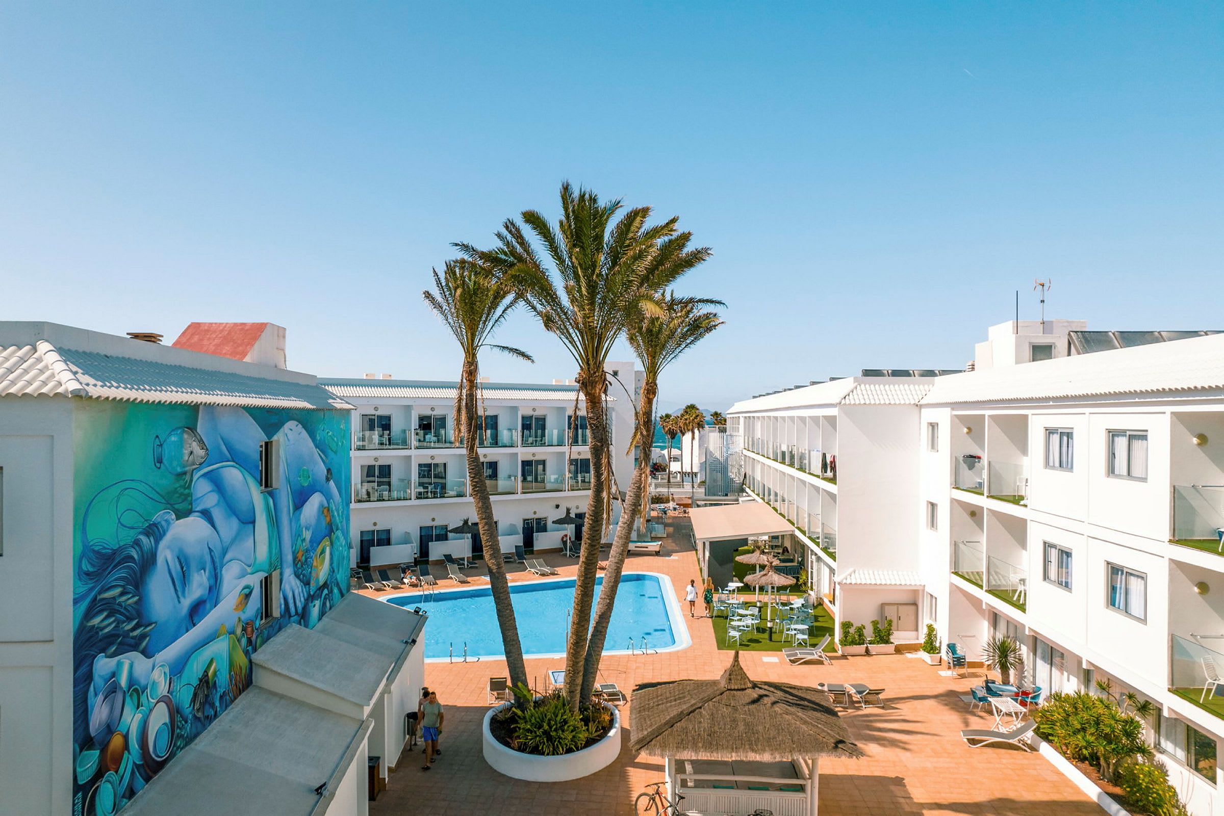 Et hotelområde med en pool i midten omgivet af hvide bygninger med altaner. Et vægmaleri ses på en af bygningerne, og der er palmer og siddepladser ved poolen. (AI-genereret tekst)