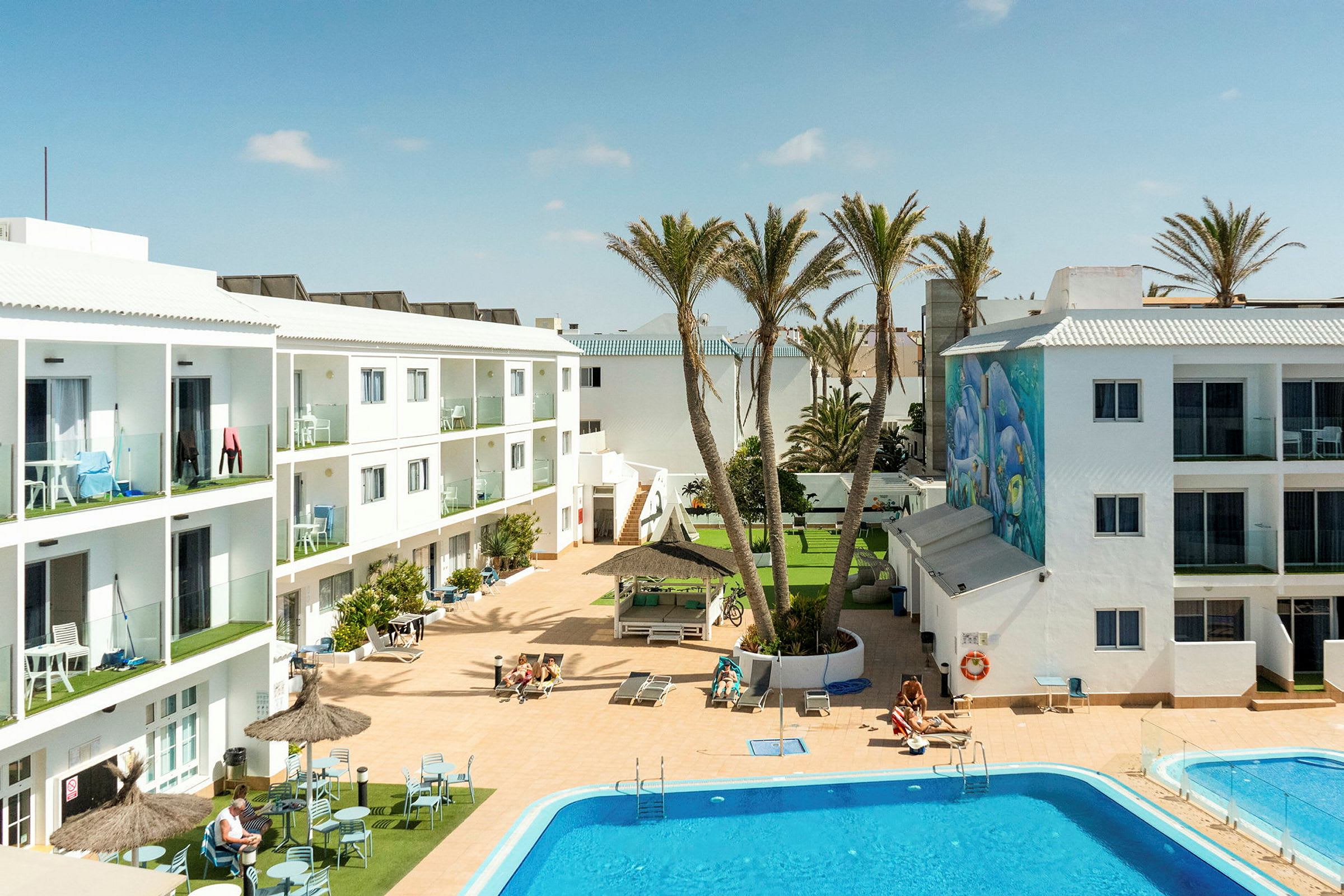 Hotelområde med hvide bygninger, en pool og liggestole. Palme og en dekoreret væg er synlig. (AI-genereret tekst)