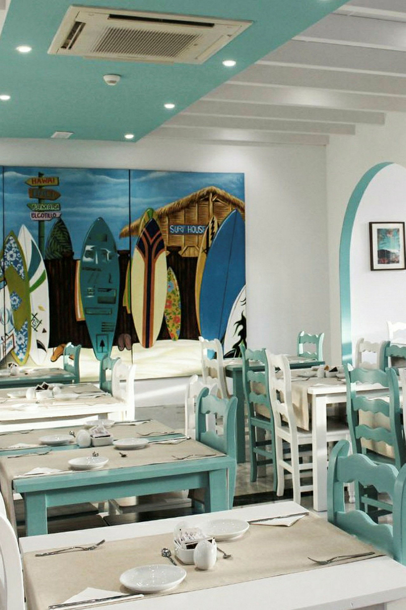 En indretning af en restaurant med turkise og hvide borde og stole. Et farverigt vægmaleri viser surfbrætter og en hytte. (AI-genereret tekst)