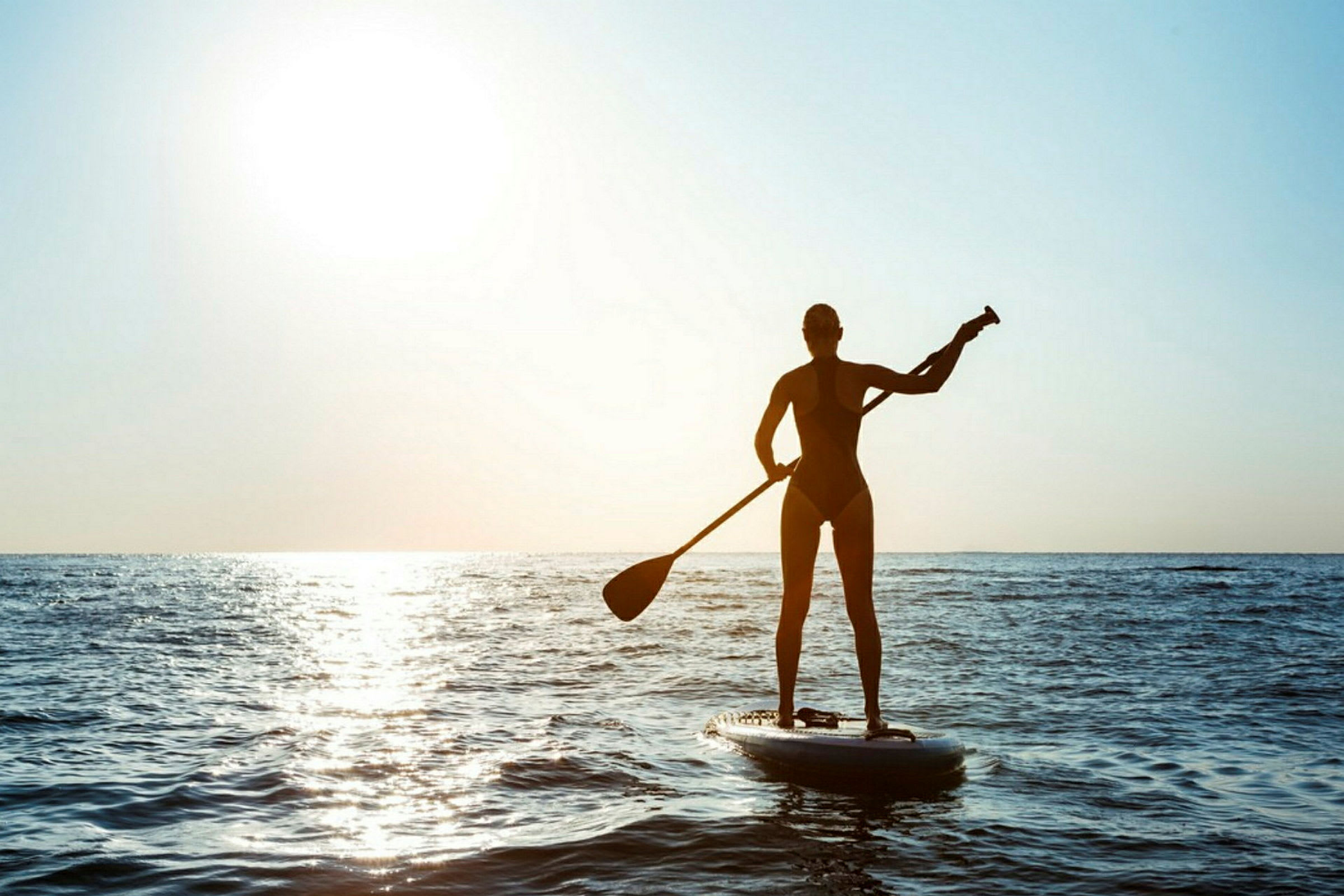 En person padler stående på et paddleboard på havet i modlys med solen i baggrunden. (AI-genereret tekst)
