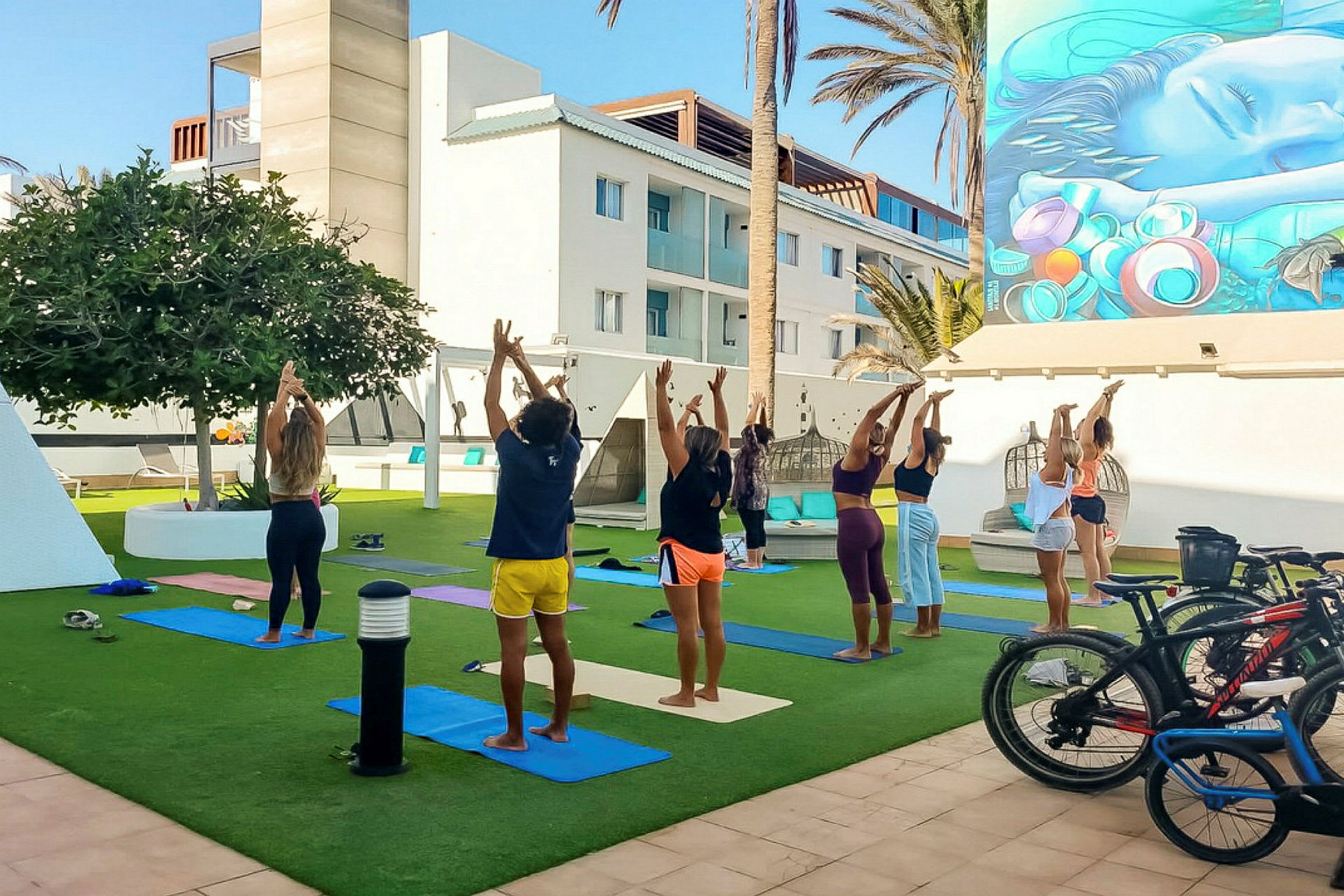En gruppe mennesker laver yoga udendørs på græs med måtter, omgivet af en moderne bygning og palmer. (AI-genereret tekst)