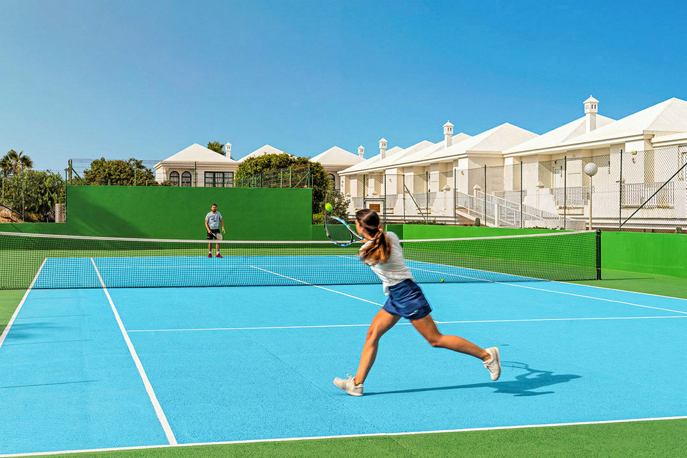 To personer spiller tennis på en udendørs tennisbane med blåt underlag og grønne omgivelser. Bag banen ses hvide bygninger og blå himmel. (AI-genereret tekst)
