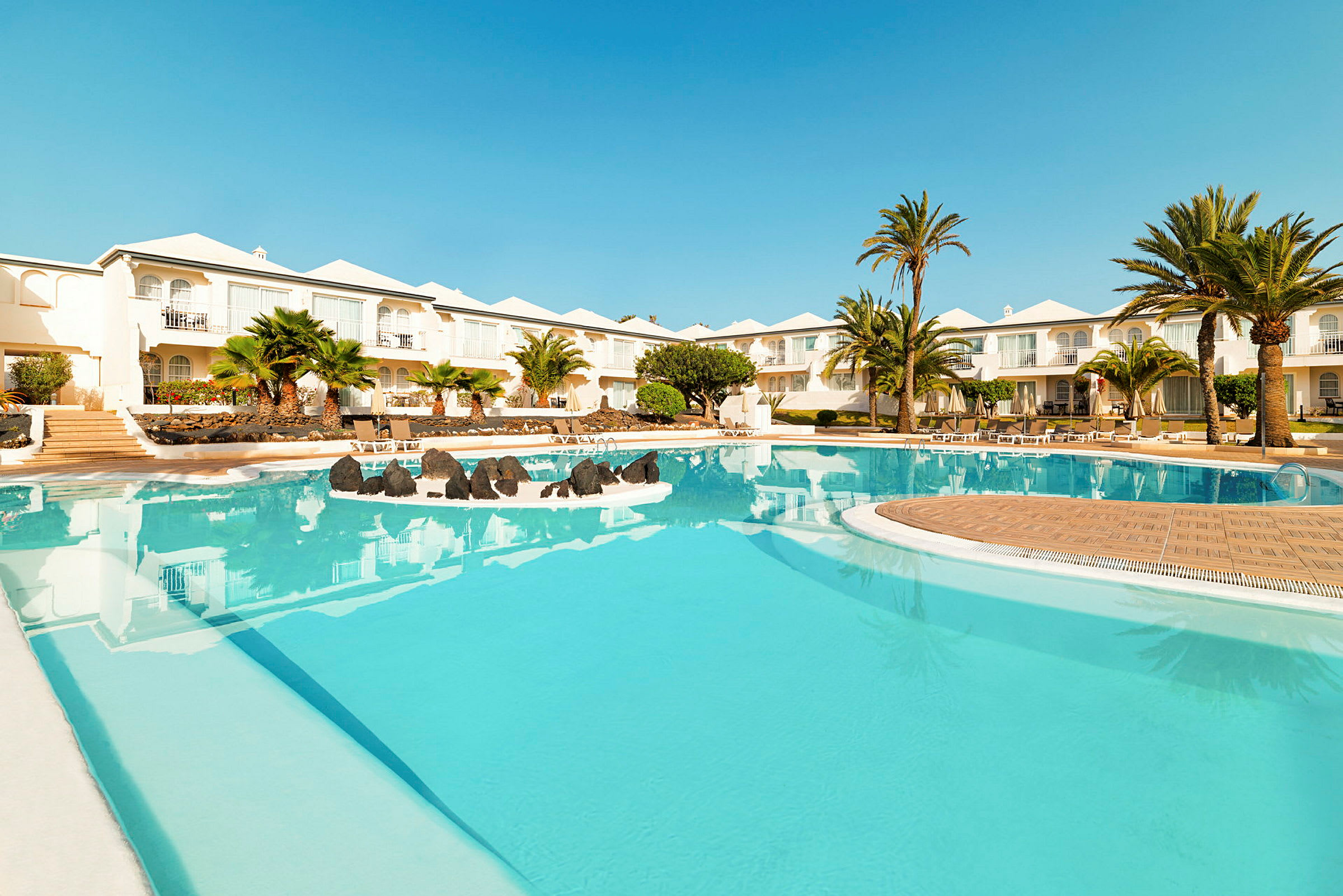 Et billede af en hotelpool med klart blåt vand, omgivet af palmetræer og hvide bygninger i baggrunden. (AI-genereret tekst)