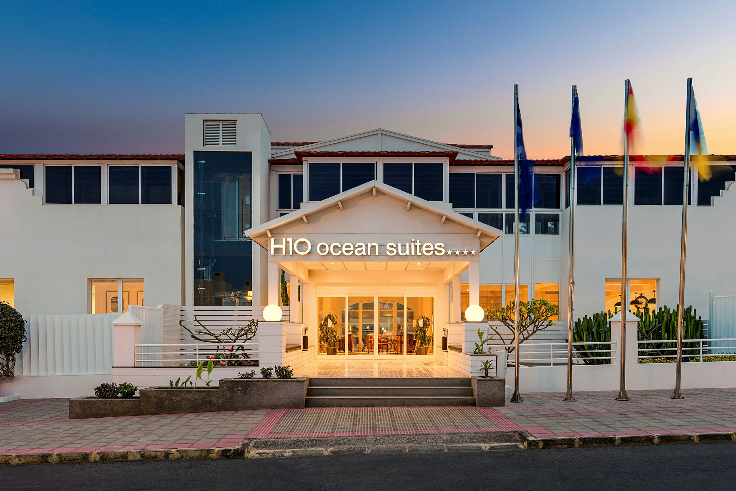 Hotelindgang med skiltet H10 Ocean Suites og flagstænger ved solnedgang. (AI-genereret tekst)