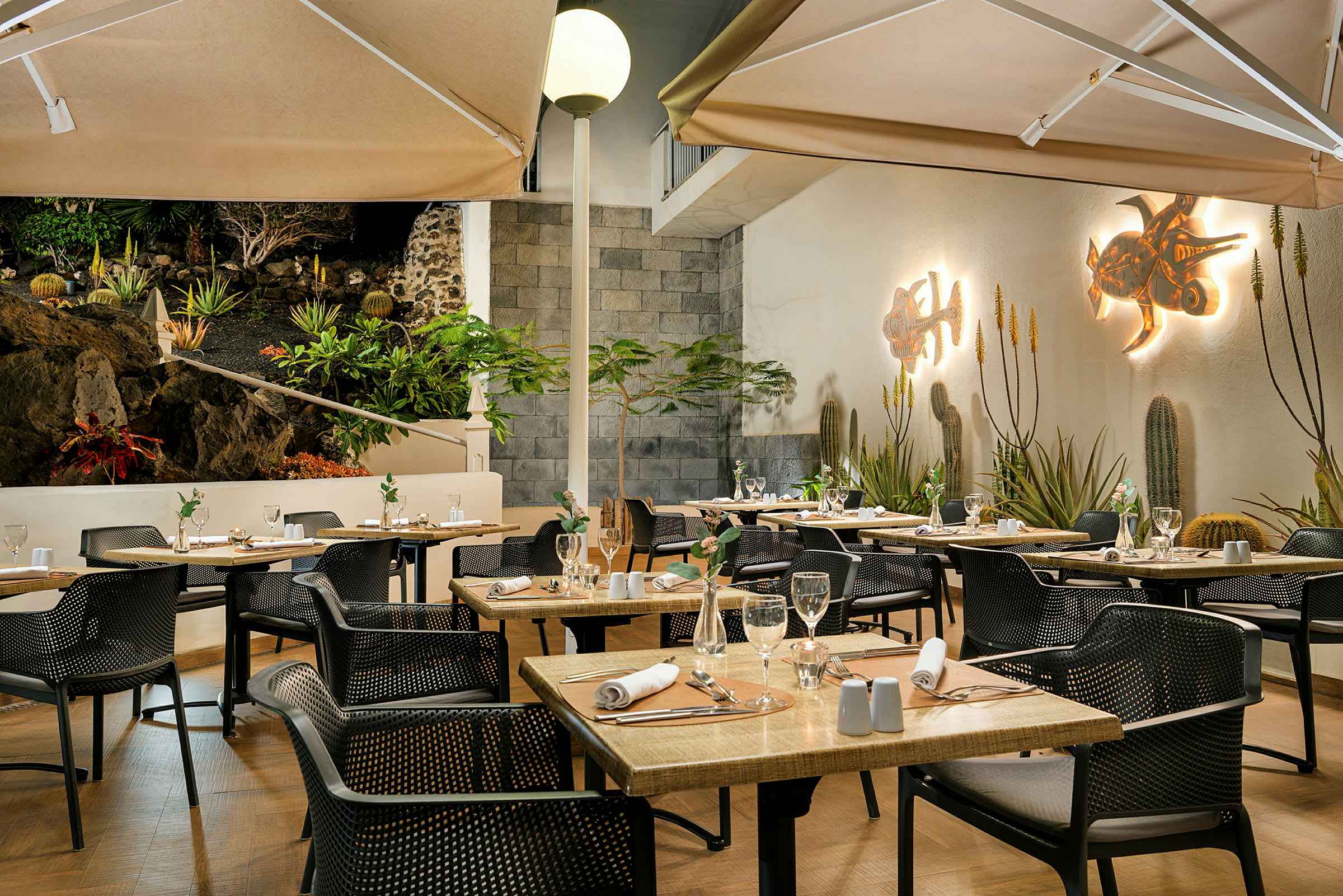 En veloplyst restaurantterrasse med dækkede borde og stole. Væggene er dekoreret med kunst, og omgivelserne har planter og en stenmur. (AI-genereret tekst)