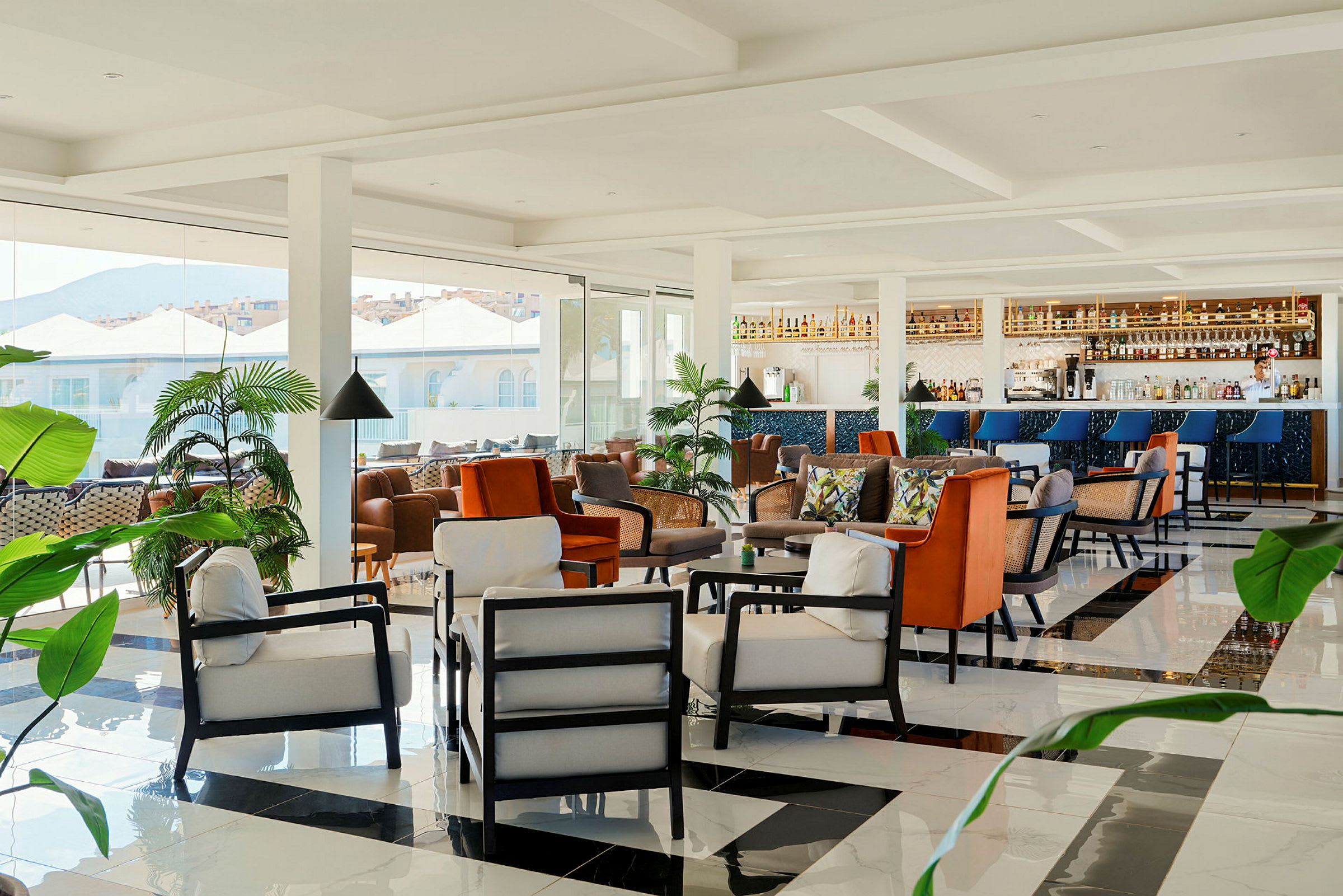 En moderne lounge med komfortable siddepladser, en bar og frodige planter samt store vinduer med udsigt til bygninger og bjerge. (AI-genereret tekst)