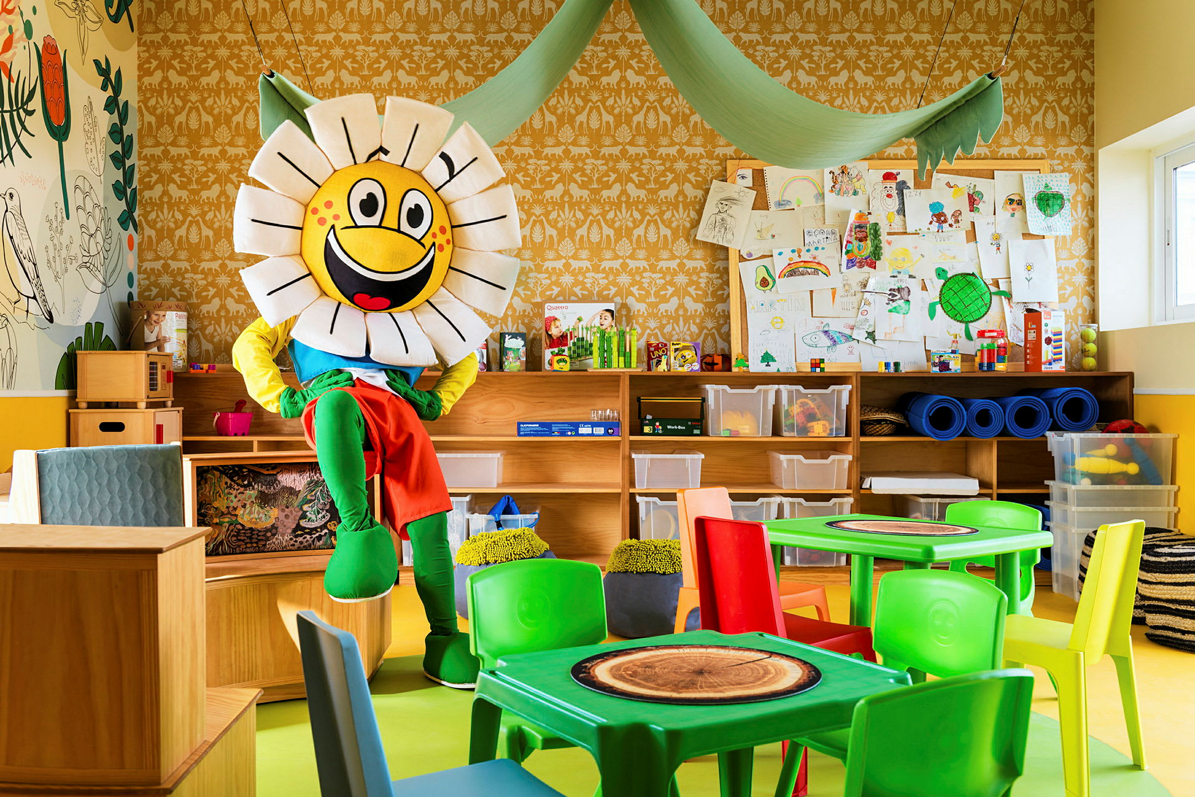 Et farverigt rum med en maskot i form af en blomst, der sidder ved møbler. Baggrunden viser hylder, opbevaringskasser og tegninger. (AI-genereret tekst)