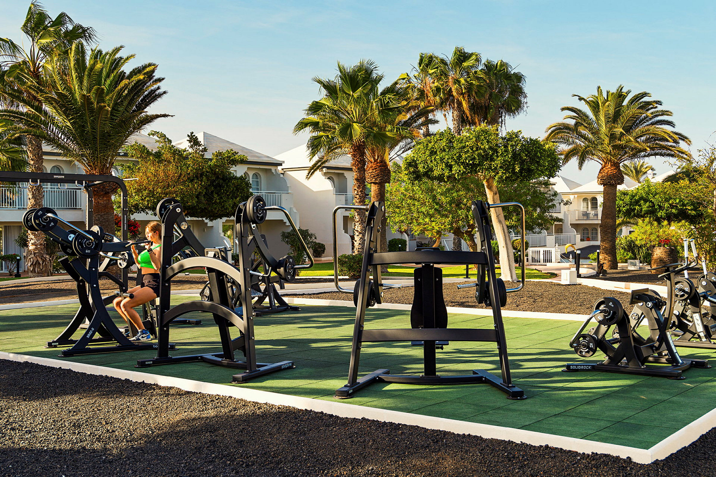 Udendørs træningsområde med fitnessudstyr placeret på grønne måtter, omgivet af palmer og bygninger. (AI-genereret tekst)