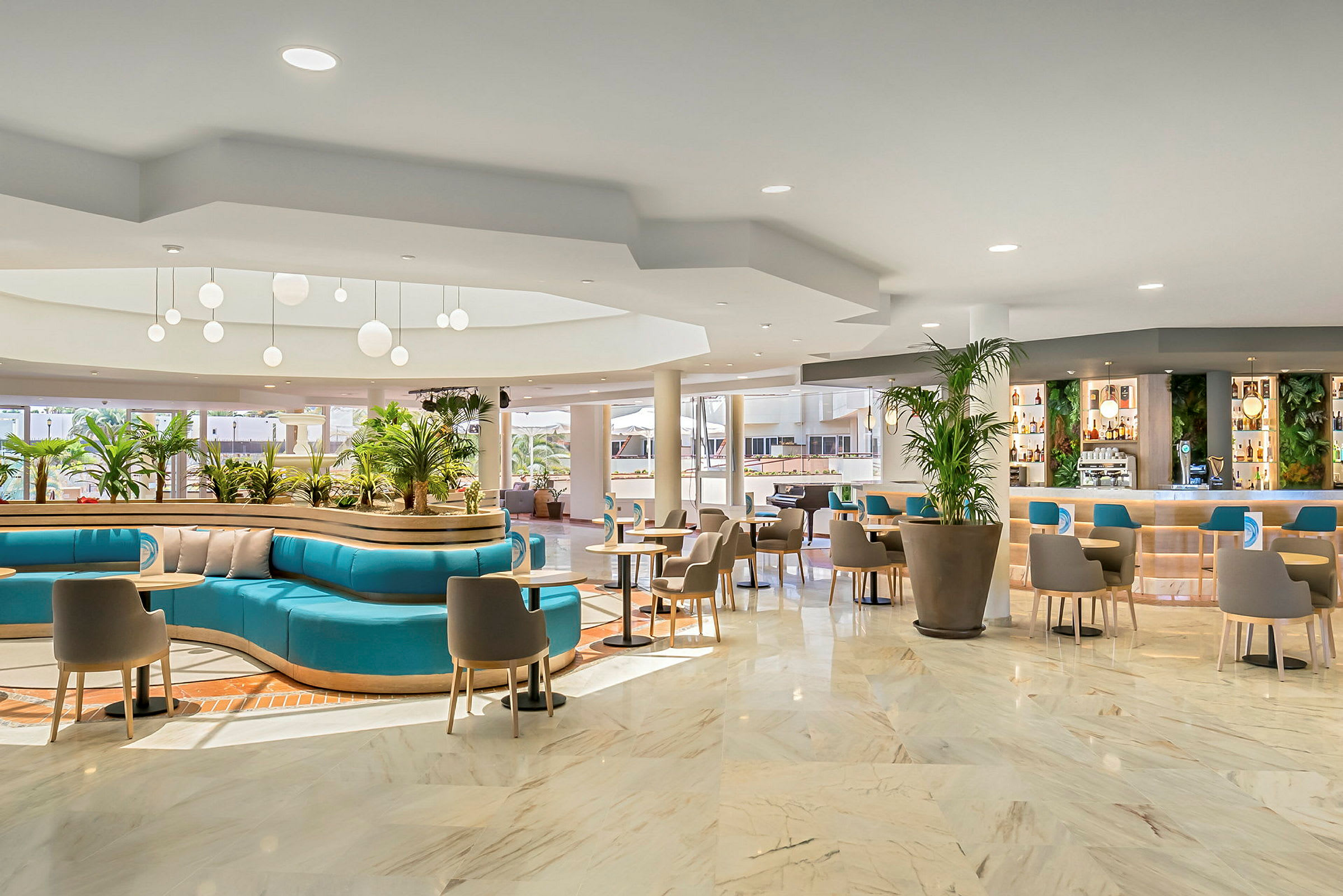 Et moderne hotelområde med siddegrupper, turkise sofaer og store potteplanter. Loftlamper hænger fra loftet, og en indbydende bar med hylder er synlig i baggrunden. (AI-genereret tekst)