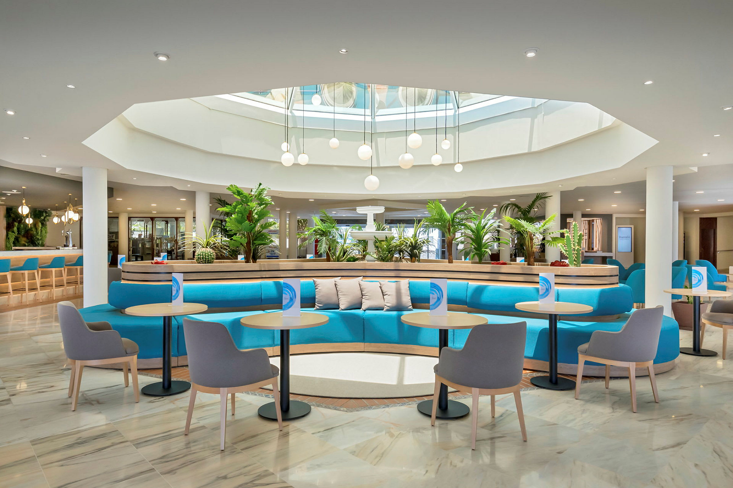 En moderne hotellobby med blå sofaer, små borde og stole. Planter pryder området, og loftsbelysning med glaskupler ses under et ovenlys. (AI-genereret tekst)