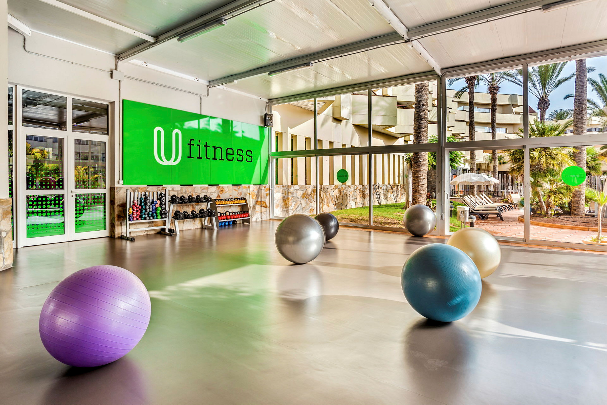 Et fitnessområde med store vinduer og træningsbolde på gulvet. Håndvægtsstativer og træningsudstyr ses ved en grøn væg mærket "fitness". (AI-genereret tekst)