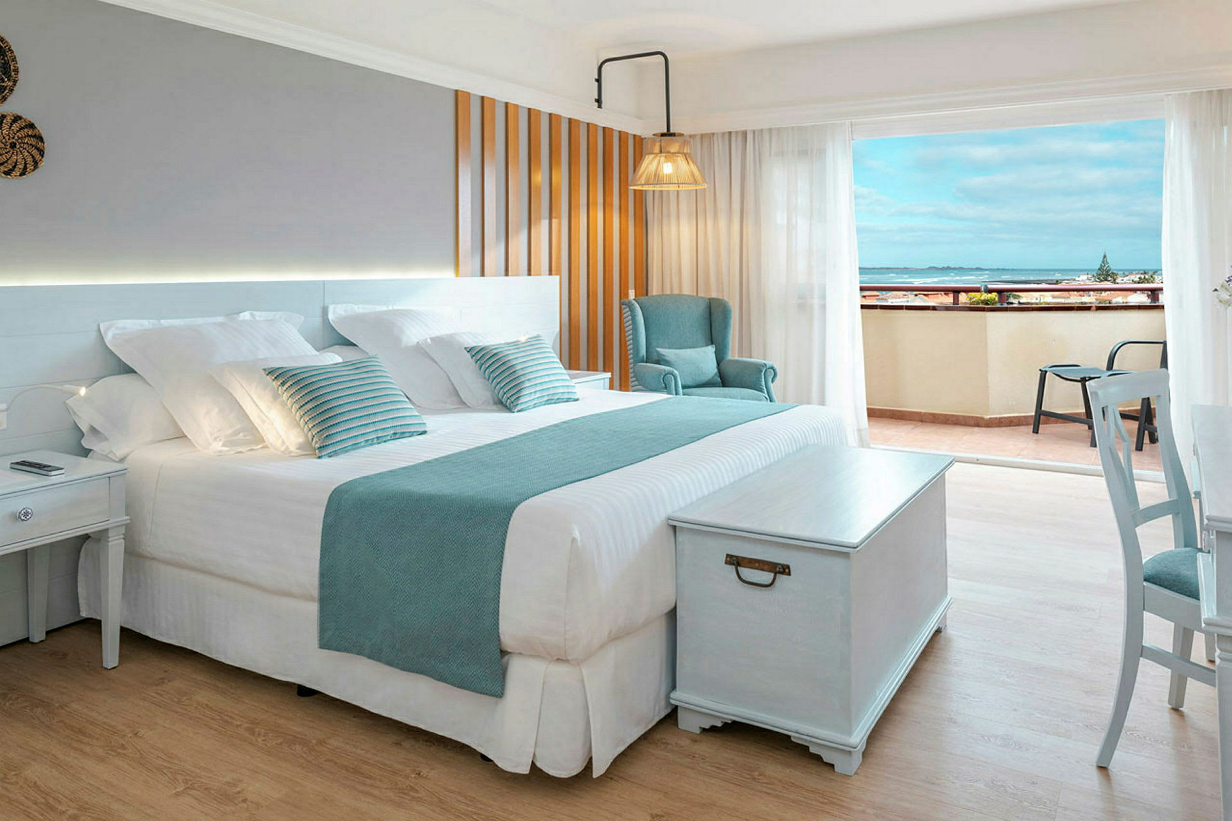 Et lyst hotelværelse med en stor dobbeltseng, stol og bord, samt en balkon med udsigt over havet. (AI-genereret tekst)