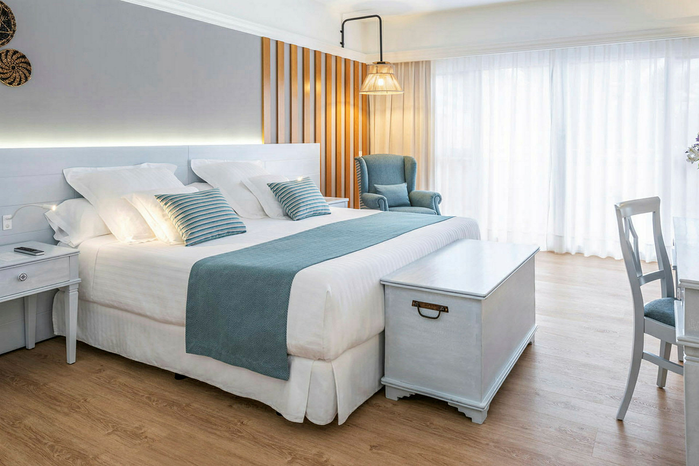 Et lyst og elegant hotelværelse med en stor seng, blå dekorationer, en lænestol og et træbord. (AI-genereret tekst)
