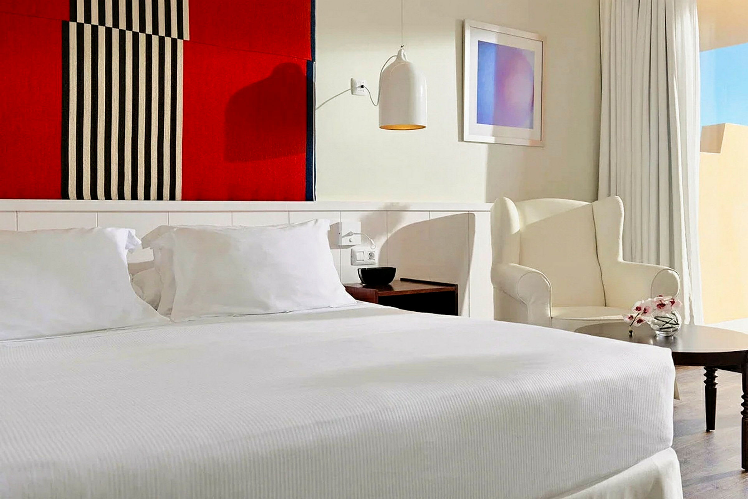 Et stilfuldt hotelværelse med en stor seng og hvidt sengetøj, en lænestol ved siden af et lille bord, samt en farverig tekstil vægdekoration over sengen. (AI-genereret tekst)