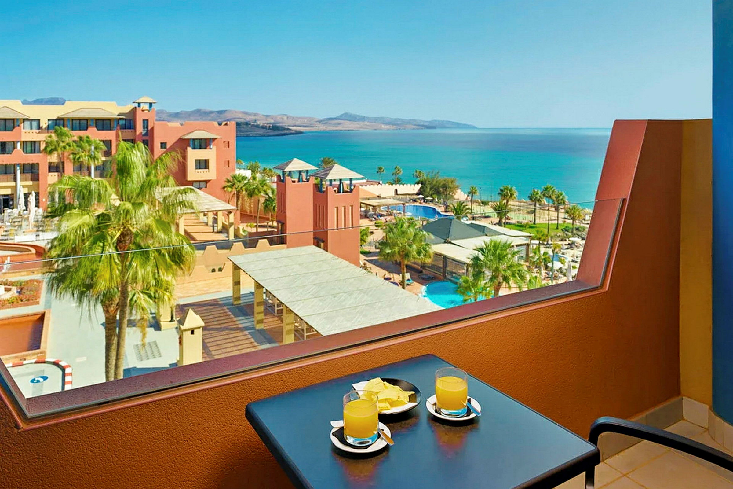 Udsigt fra en balkon med et bord med to glas juice og nogle frugtstykker. I baggrunden ses en resort med pools, palmer og havet. (AI-genereret tekst)