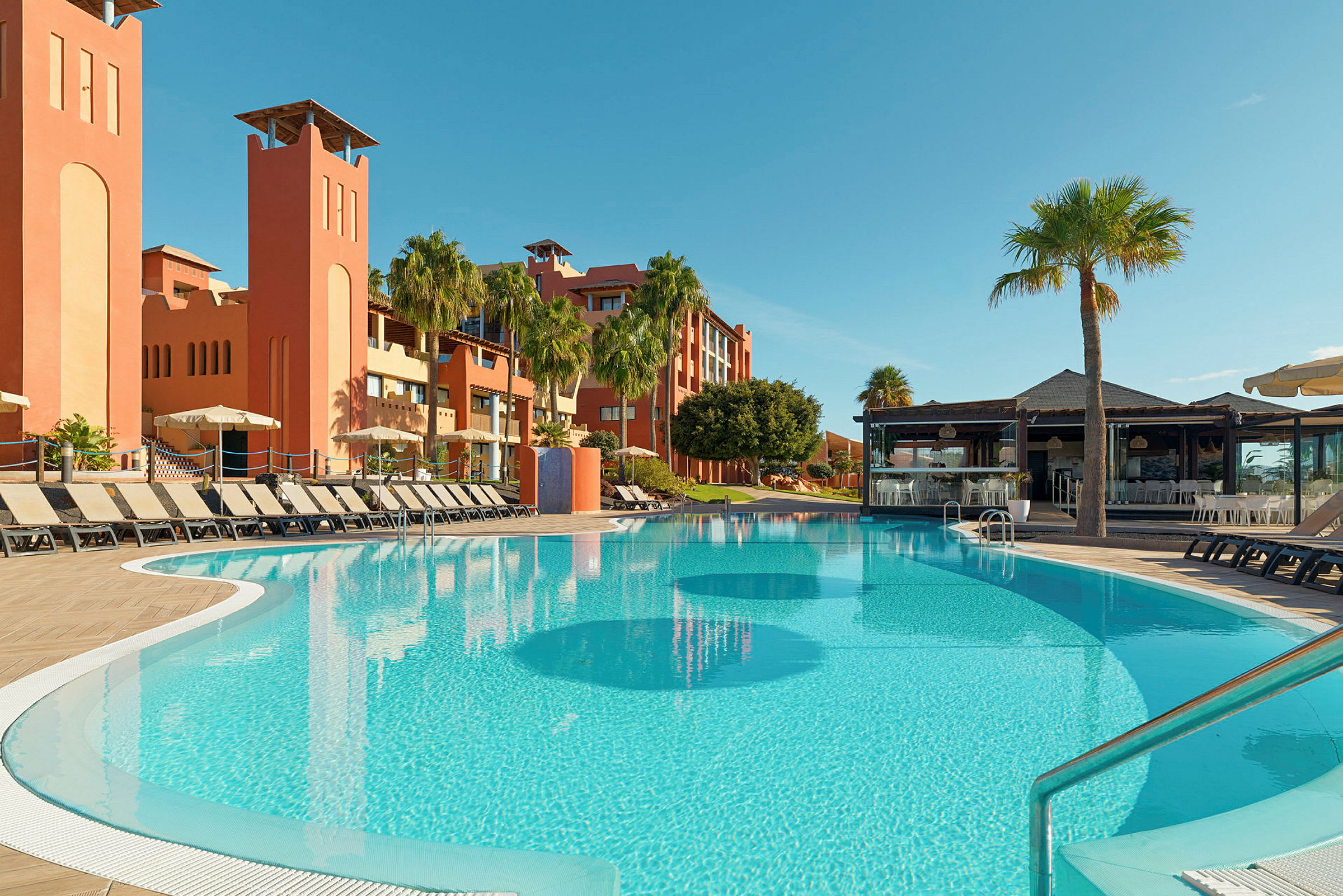 Udendørs pool med liggestole i et resortområde, omgivet af palmer og orange bygninger. (AI-genereret tekst)