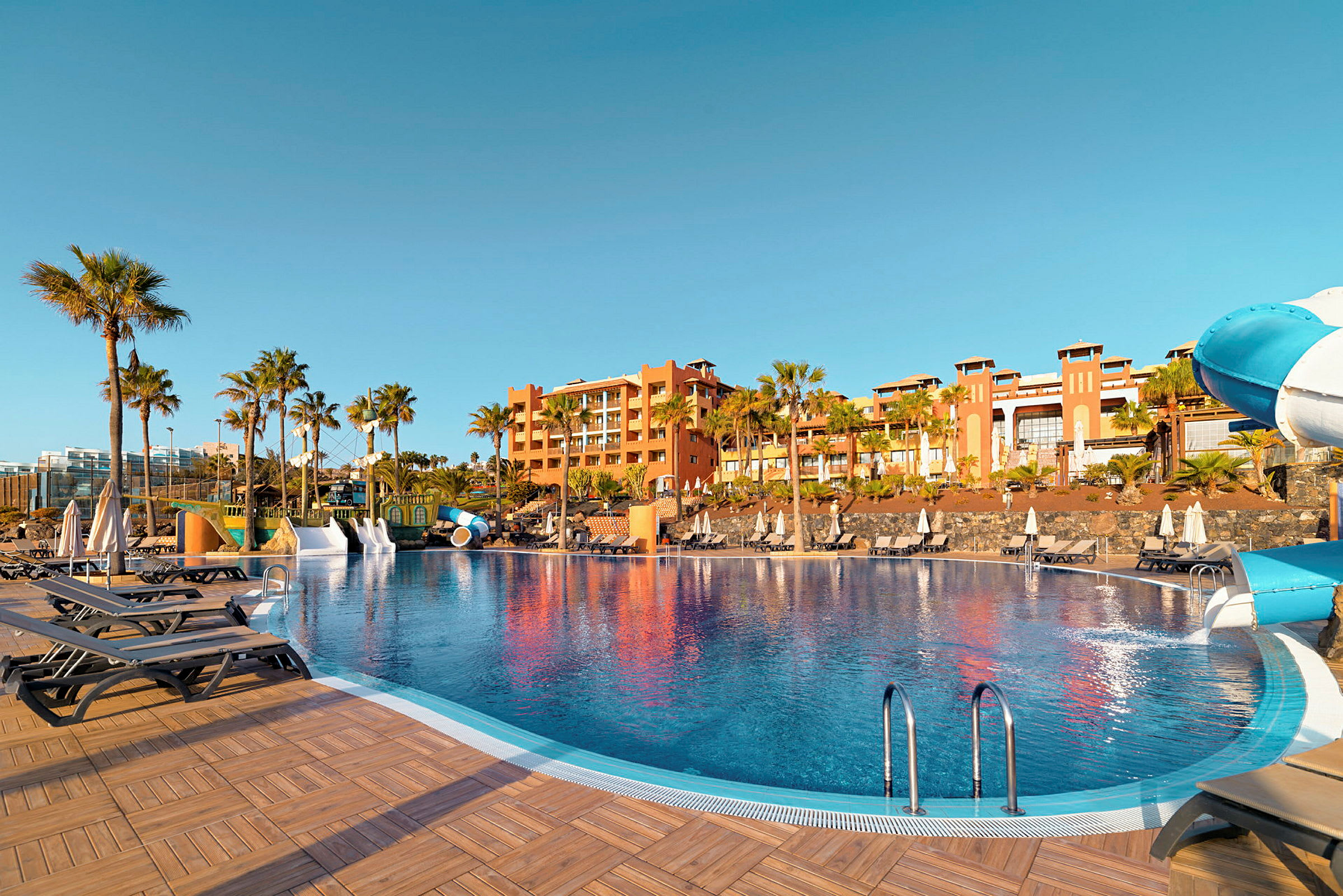 En udendørs swimmingpool med liggestole og parasoller ved et hotel omgivet af palmer. (AI-genereret tekst)