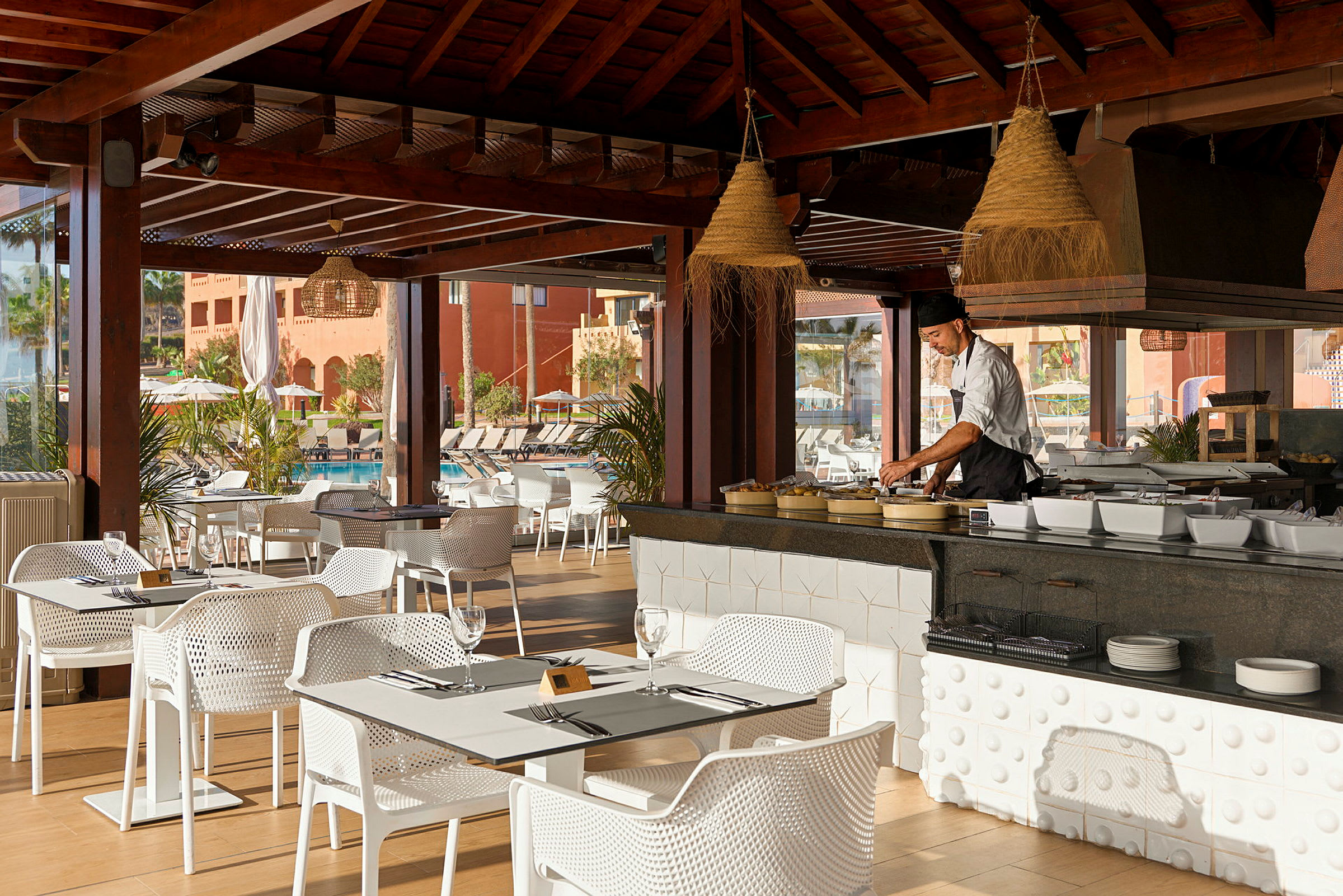 En restaurant med træmøbler på en terrasse. Bordene og stolene er hvide, og en kok arbejder ved madstationsområdet i midten. (AI-genereret tekst)