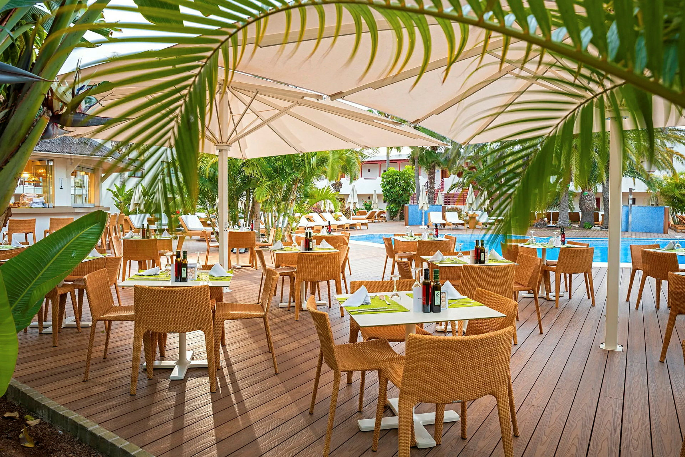 Udendørsrestaurant med trædæk, borde med duge og stole, omgivet af grønne områder og en swimmingpool. (AI-genereret tekst)