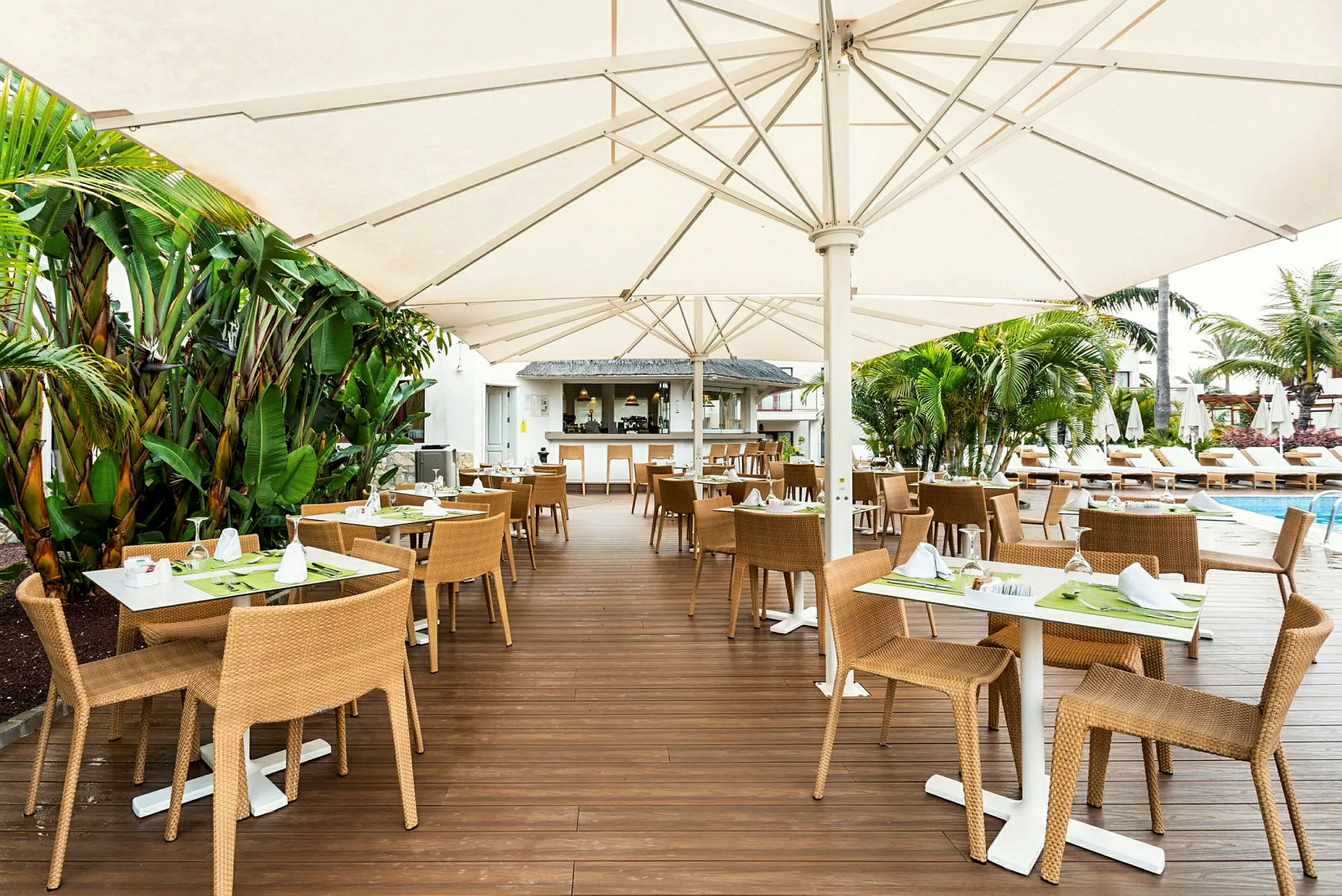 Udendørs serveringsområde med borde og stole placeret på en træterrasse, omgivet af grønne planter og med solsenge ved en pool i baggrunden. (AI-genereret tekst)