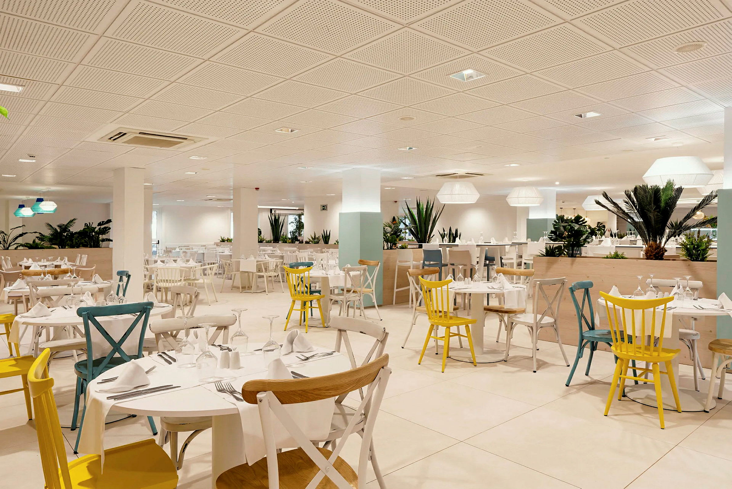 Indretningen af en lys restaurant med mange borde og stole i forskellige farver, dekoreret med grønne planter. (AI-genereret tekst)