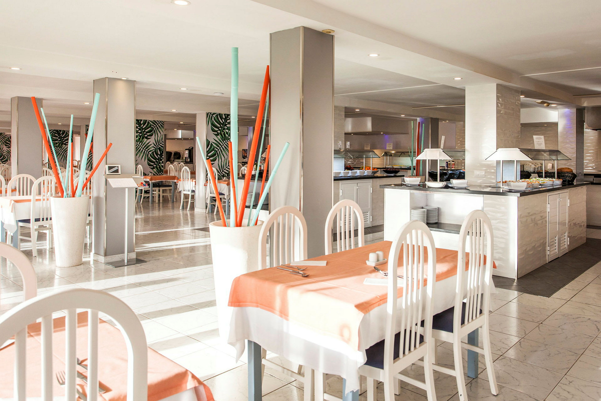 En restaurant med lyse møbler, borde dækket med duge, og dekorative elementer som farvestrålende stavinstallationer. (AI-genereret tekst)