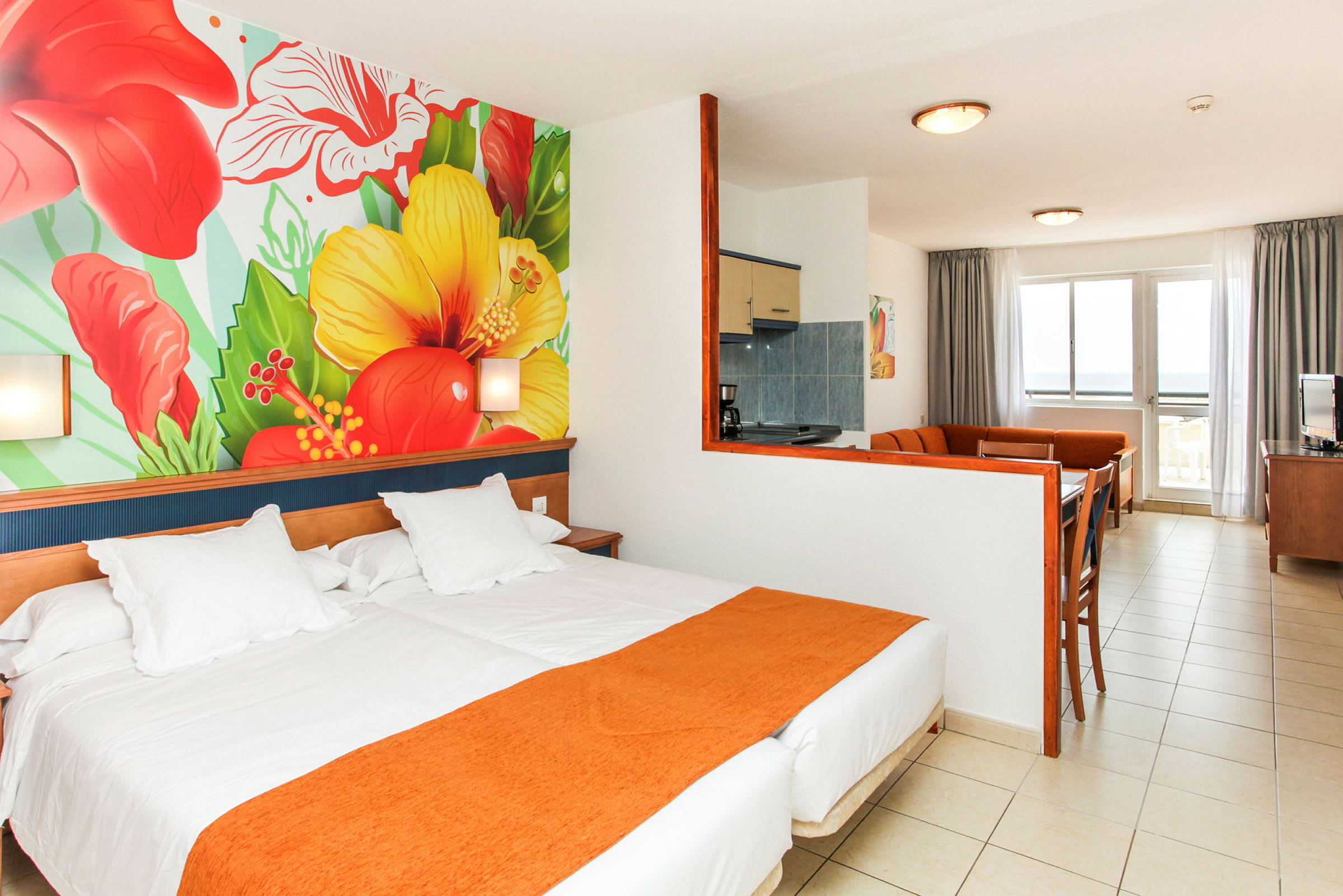 Hotelværelse med seng, farverigt vægmaleri af blomster og siddeområde med køkken. (AI-genereret tekst)