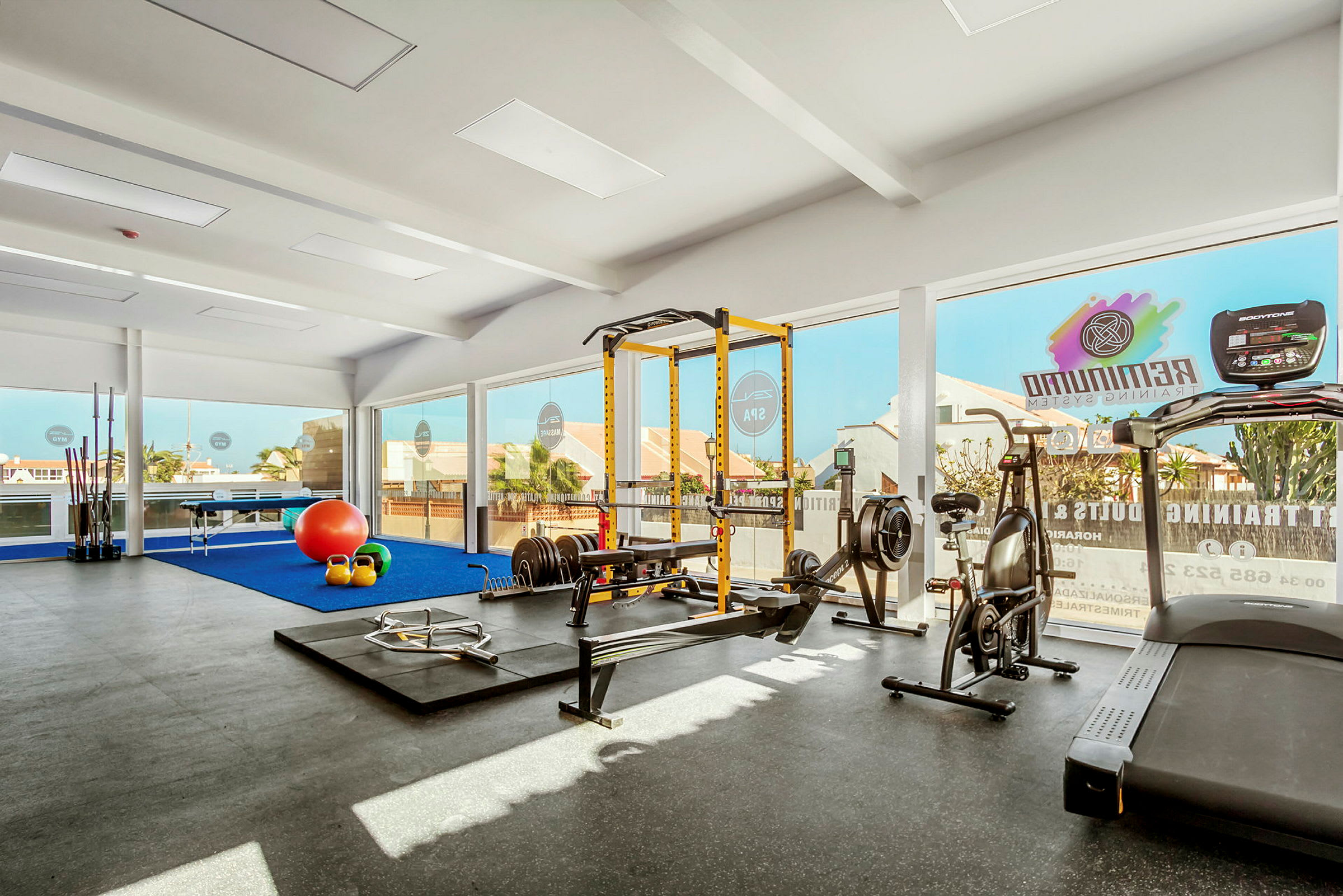 Et fitnesscenter med udstyr, herunder løbebånd, romaskine, vægte og et træningsstativ. Store vinduer lukker dagslys ind. (AI-genereret tekst)