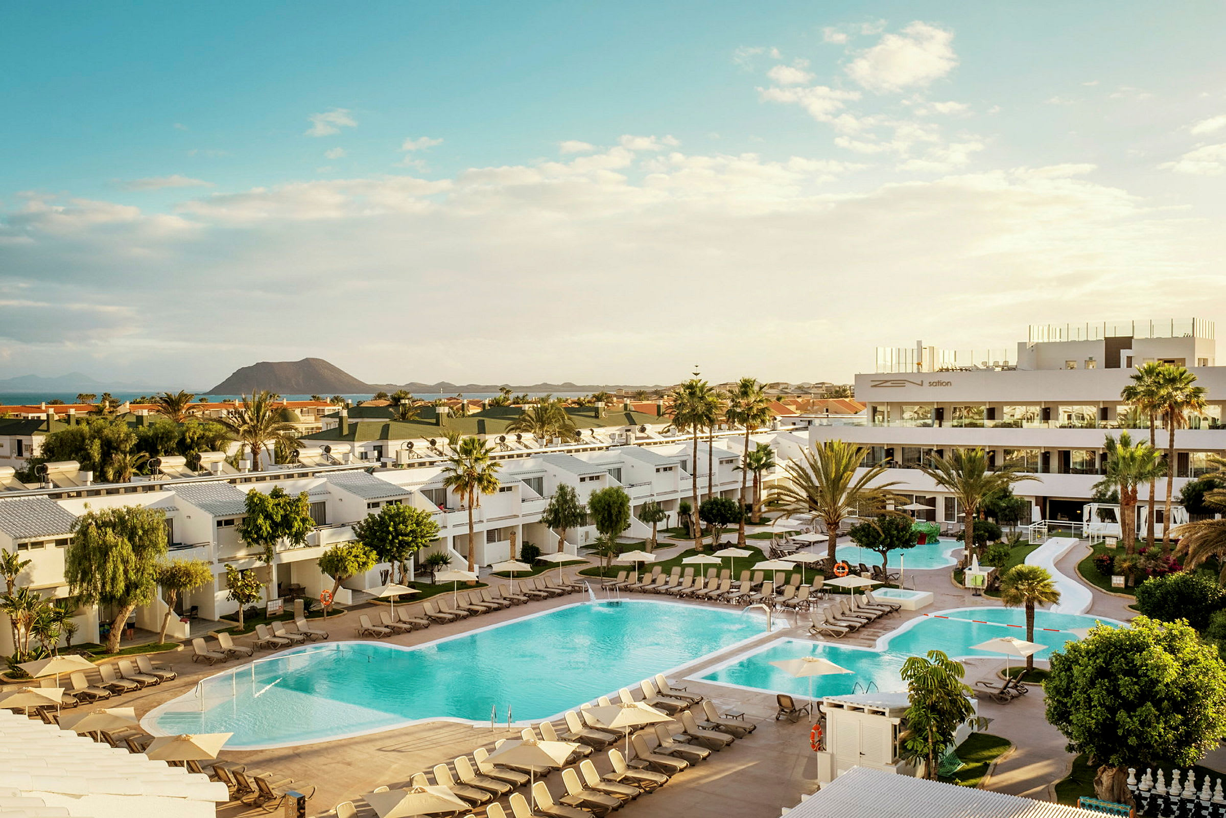 Billede af et hotelområde med pools, liggestole og omkringliggende bygninger samt en vulkan i baggrunden. (AI-genereret tekst)