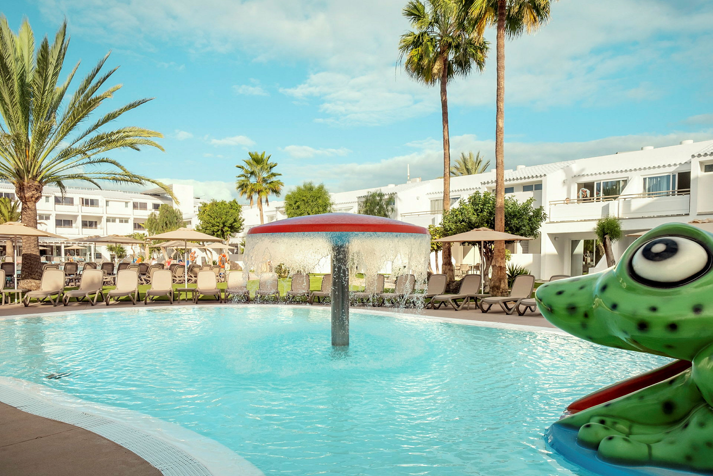 En pool med en svampeformet springvand i midten, omgivet af liggestole og parasoller. I forgrunden ses en figur formet som en grøn frø. (AI-genereret tekst)