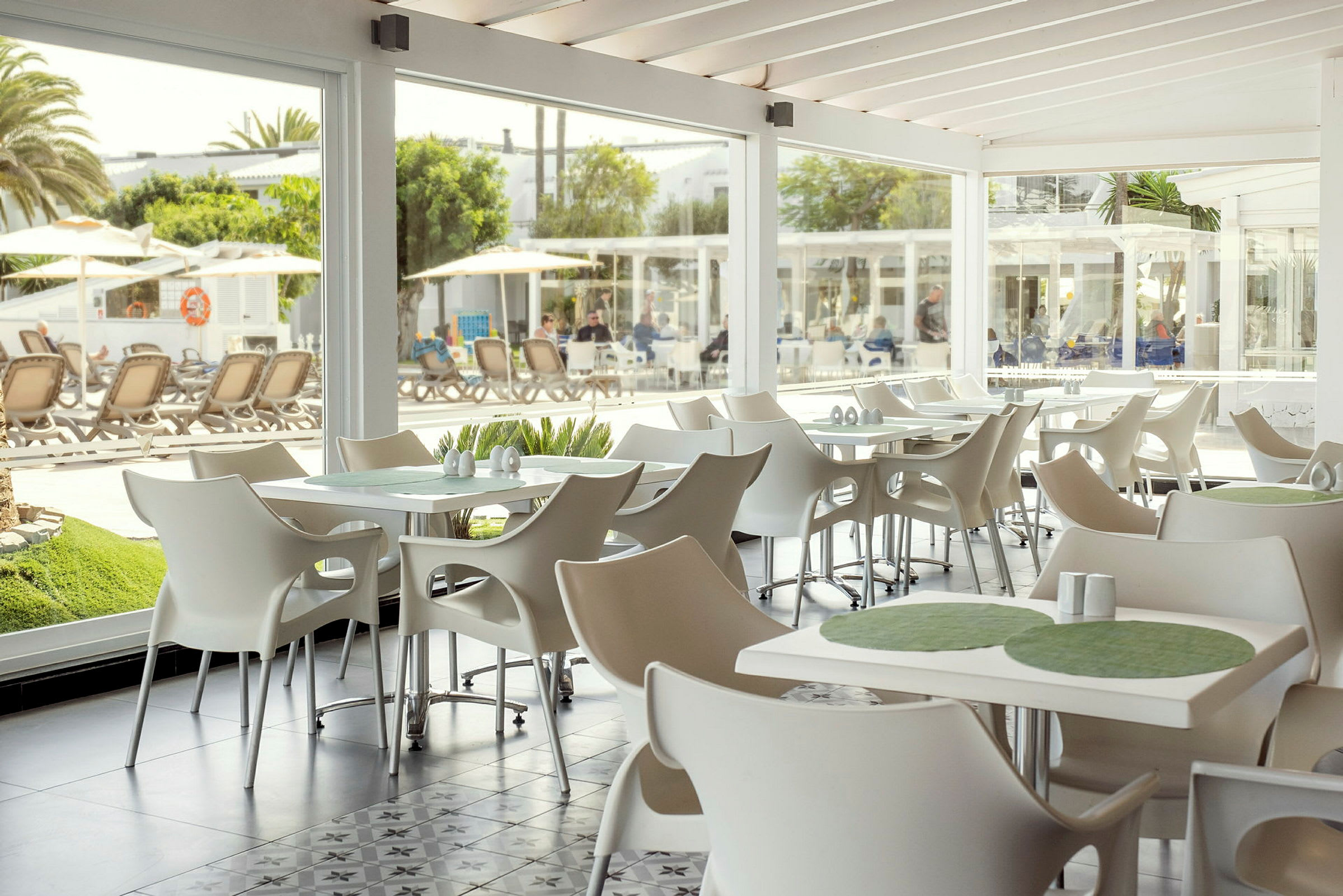 En lys og moderne indrettet café med hvide stole og borde, med udsigt til udendørs poolområde gennem store glasvinduer. (AI-genereret tekst)