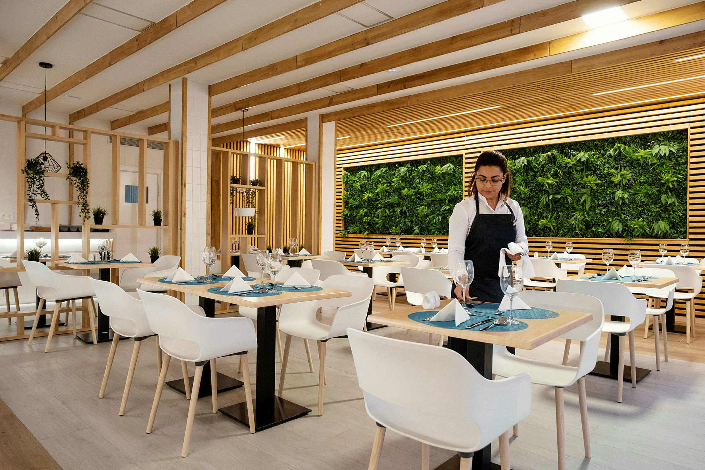 En restaurant med moderne indretning med hvide stole, dækkede borde og en væg dekoreret med planter. (AI-genereret tekst)