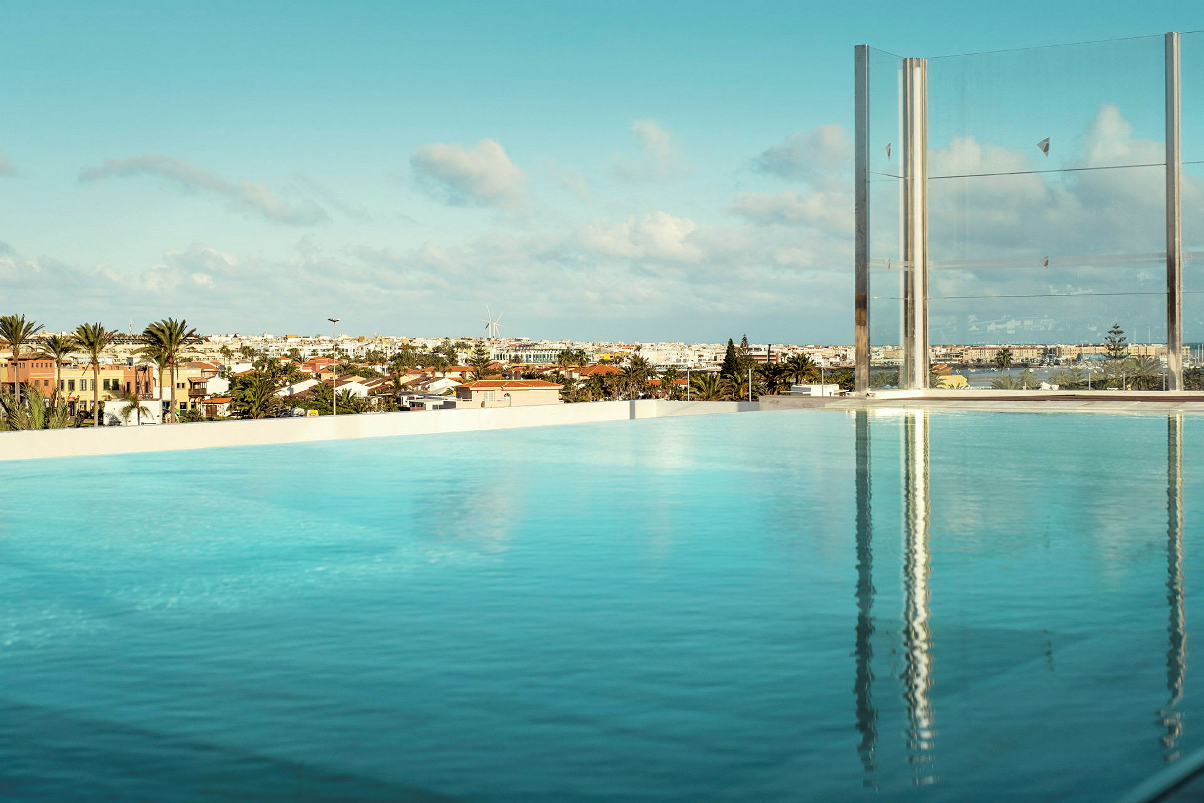 En infinity-pool med udsigt over byhuse, palmer og en vindmølle under en blå himmel. (AI-genereret tekst)